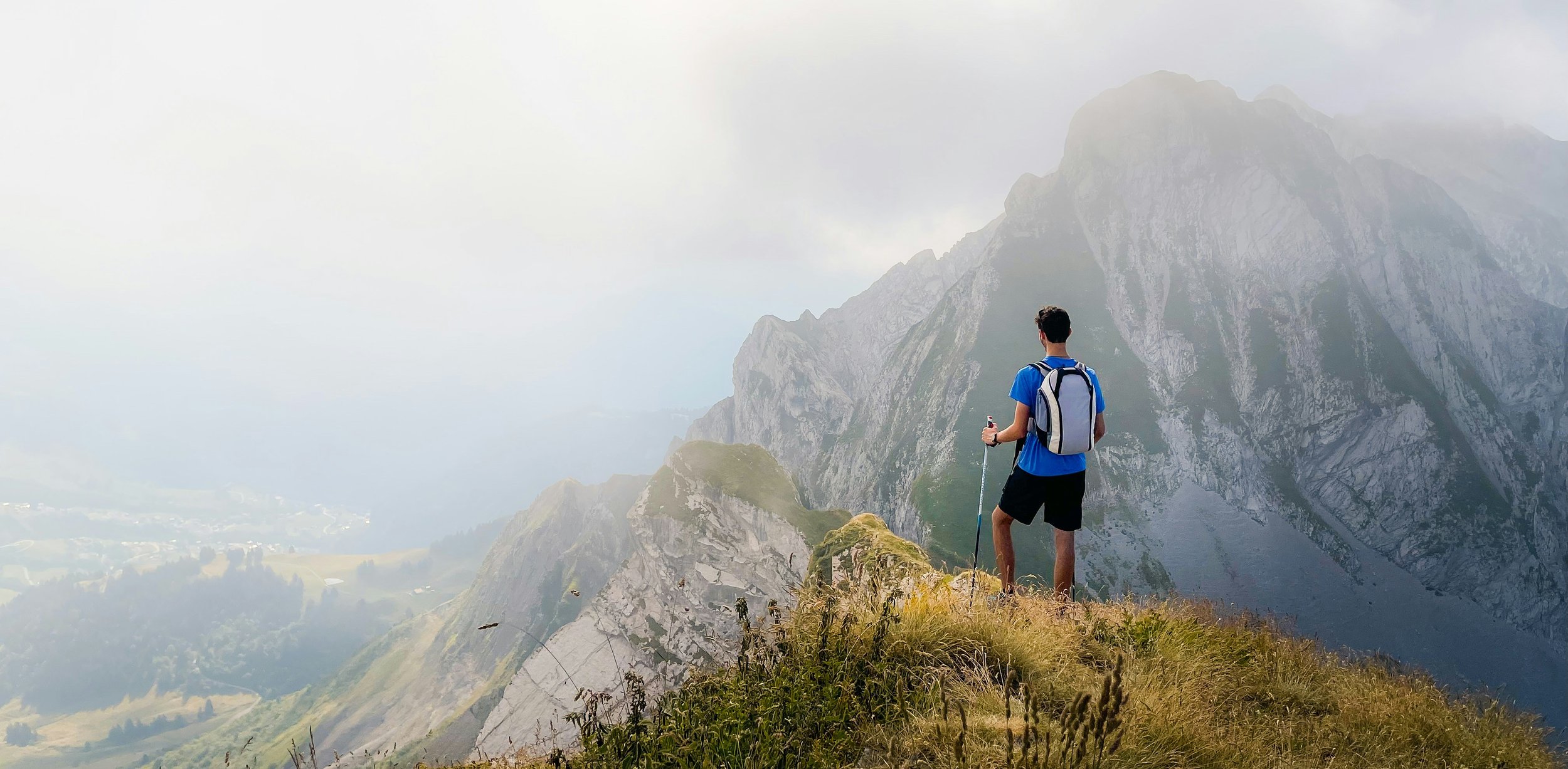 A man with a backpack and hiking poles standing on a grassy mountain trail, overlooking a mountain range with rocky peaks, some clouds, and a valley below.