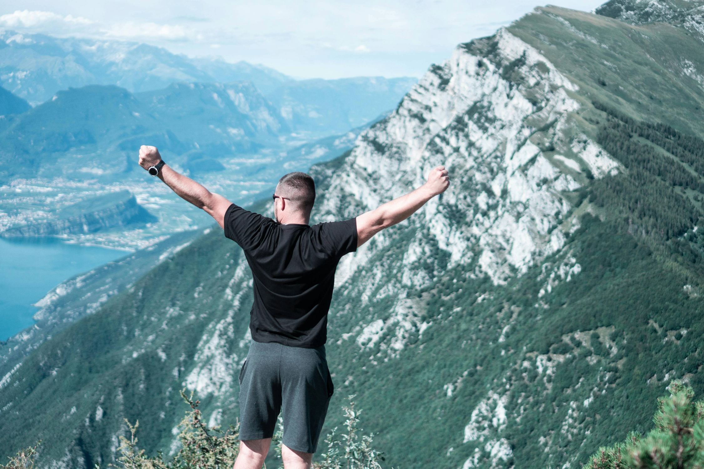 A man stands on a mountain peak with arms raised towards the sky, overlooking a scenic view of mountains and a lake in the distance.