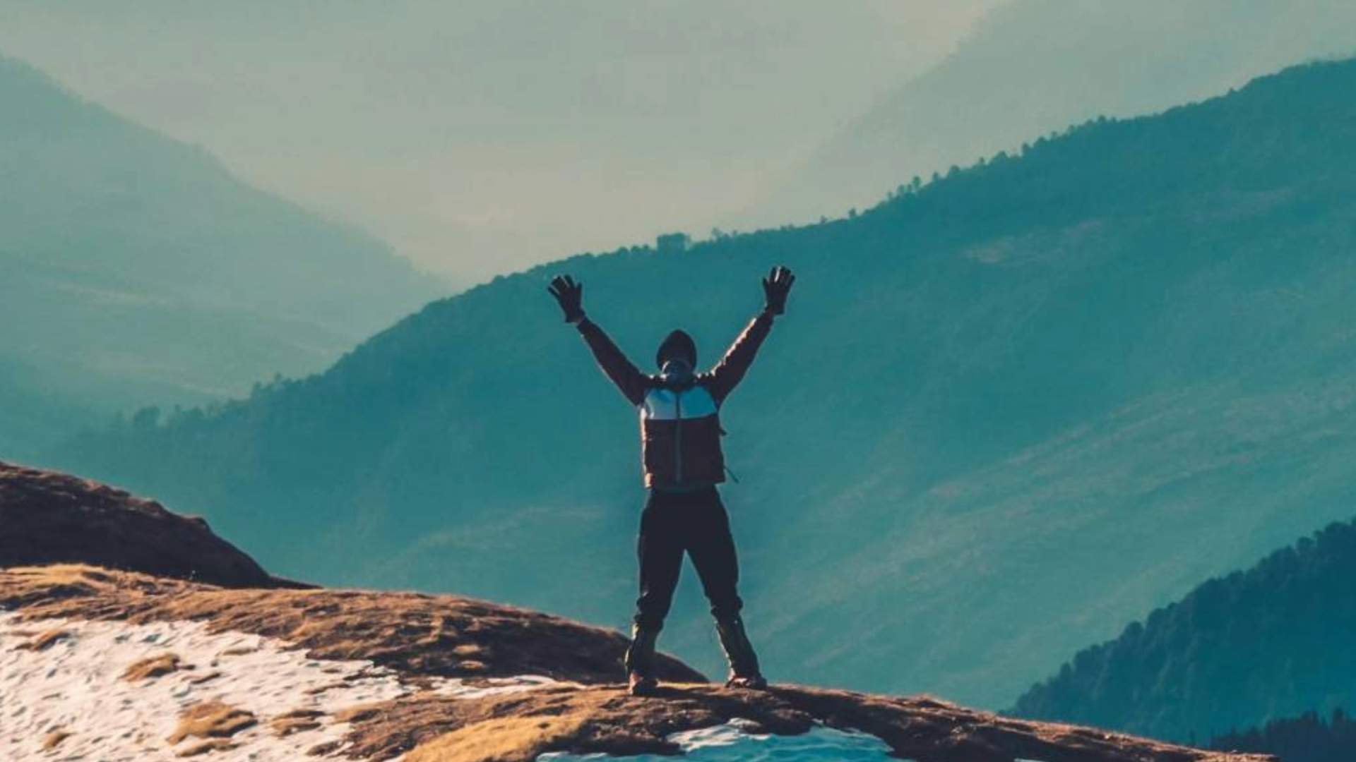 Person standing on a rocky mountainside with arms raised, overlooking green valleys and mountain ranges.