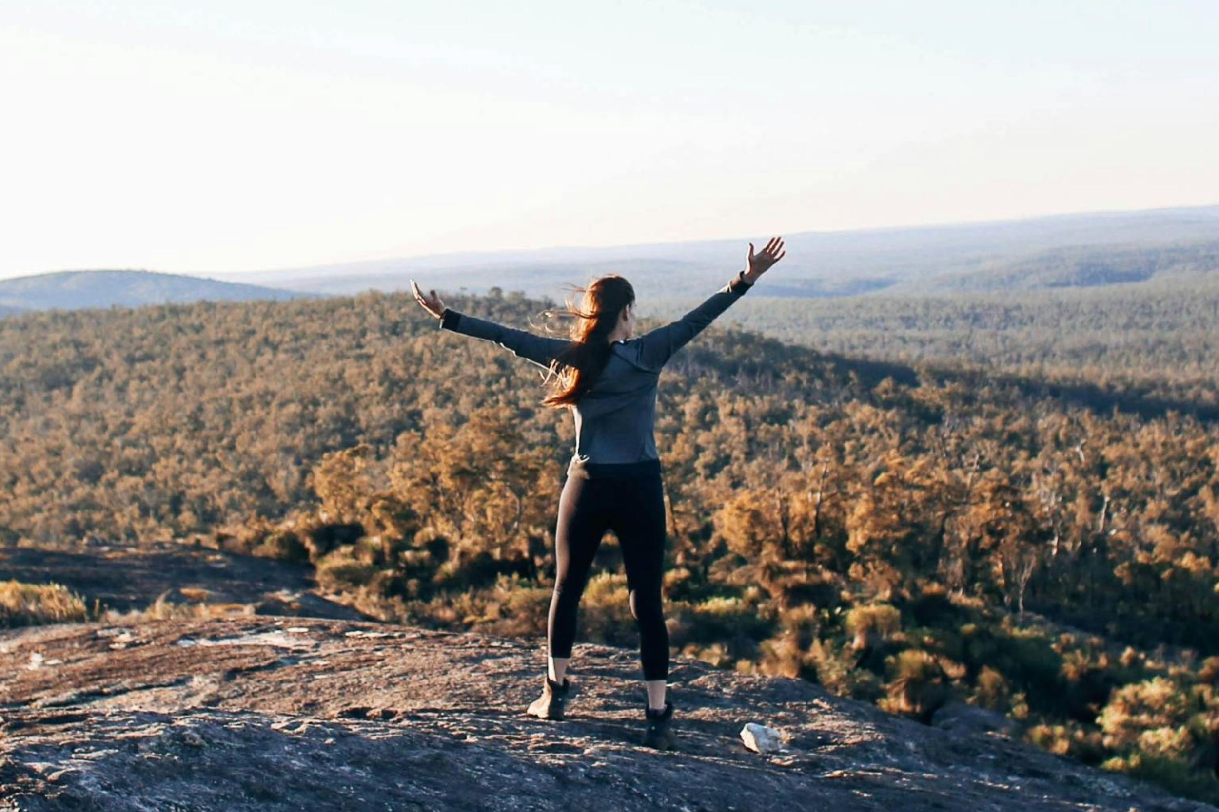 A woman standing on a rocky ledge with her arms raised, overlooking a vast forested landscape under a clear sky.