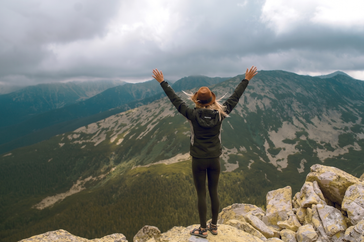 A woman standing on rocky terrain with her arms raised, overlooking a mountain range with cloudy skies.