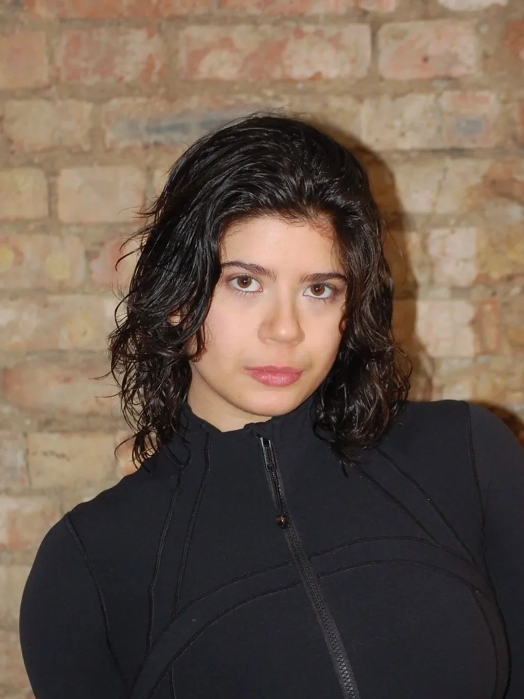 A young woman with medium-length curly dark hair and light skin, wearing a black zip-up top, standing in front of a brick wall.