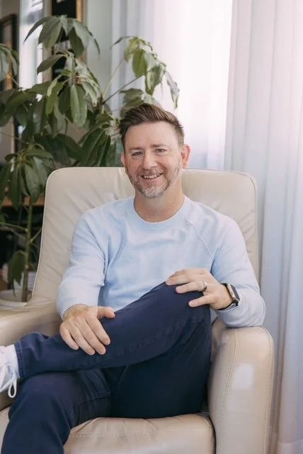 A man with short hair and a beard smiling while sitting in a cream-colored armchair in a bright room with large plants and sheer curtains.