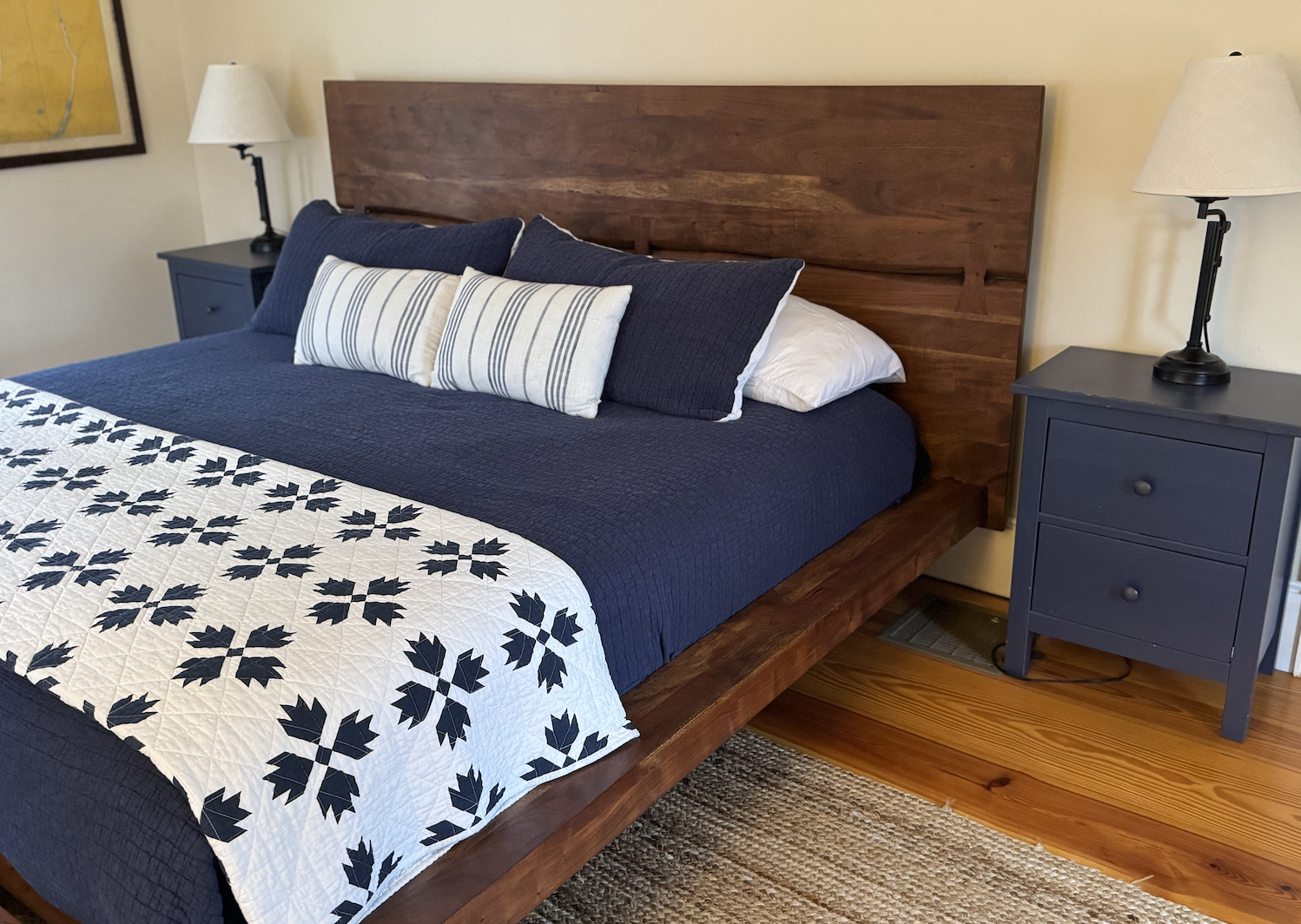 A neatly made bed with a dark blue quilt, white pillows with blue and white striped pillows in front, a wooden headboard, and two blue nightstands with lamps in a bedroom.