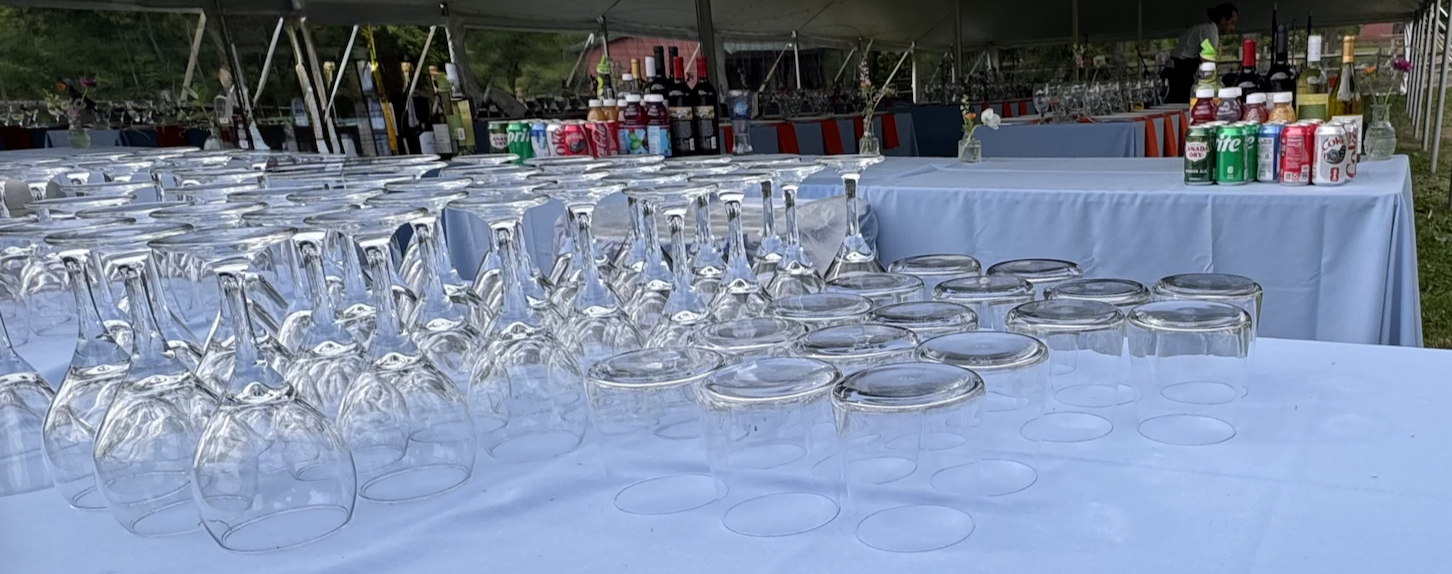 Rows of upside-down wine glasses and empty glasses on a table at a catered outdoor event with drinks in cans and bottles, and a person in the background preparing drinks.