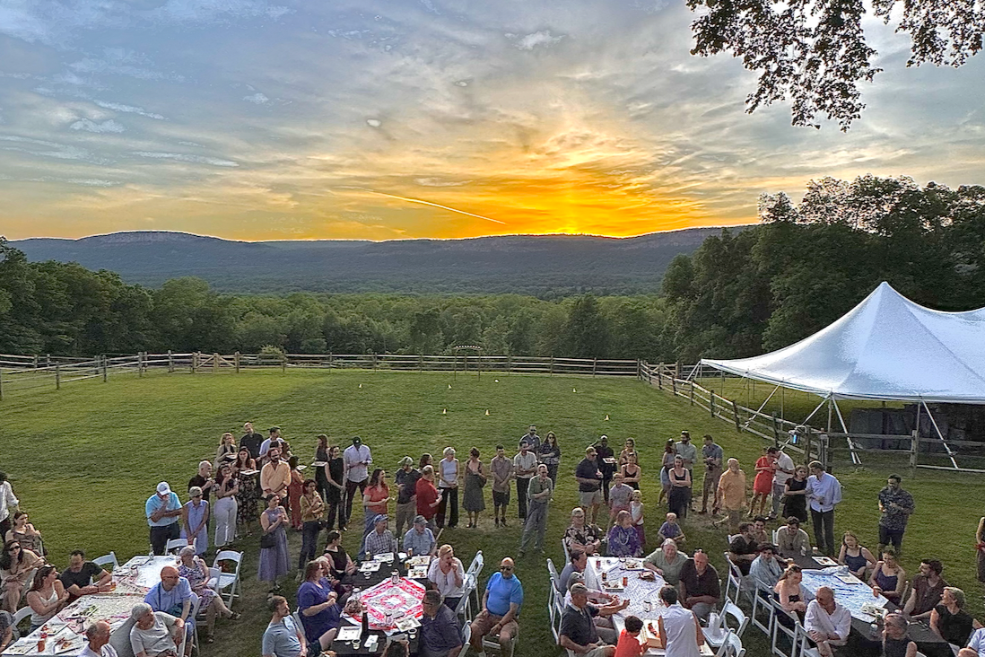 A large cocktail gathering at sunset, with tables, chairs, and a tent set up in a grassy field surrounded by trees and the Shawangunk mountains.