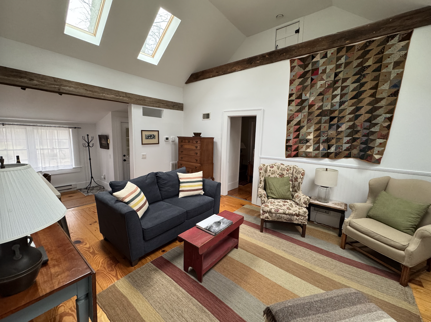 Living room with hardwood floors, striped area rug, dark blue sofa with striped pillows, floral armchair with green pillow, beige armchair with green pillow, wooden coffee table with book, side table with lamp, tall wooden dresser, wall tapestry, skylights in ceiling, window with curtains, coat rack near window.