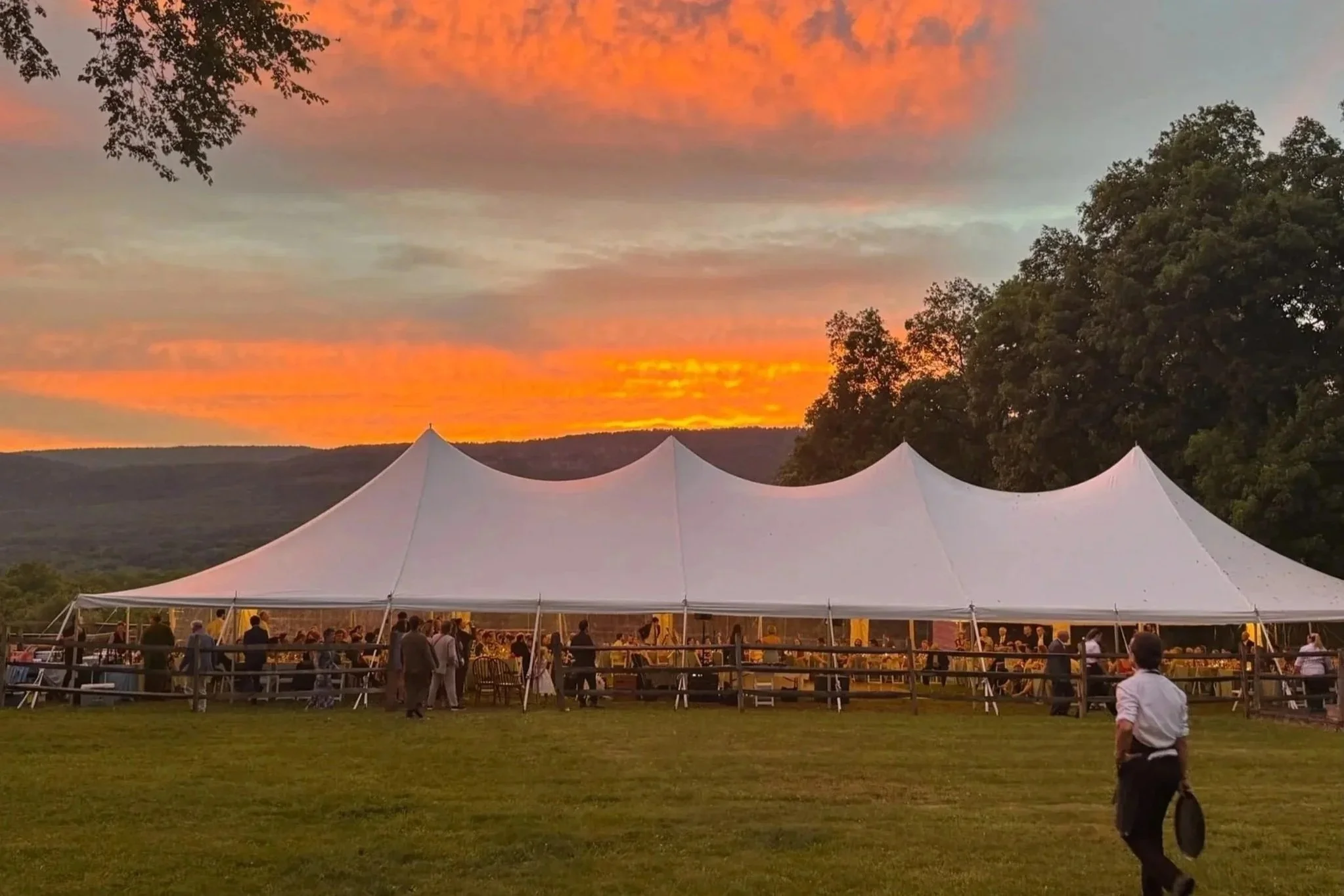 People gathered under a large white tent during sunset, with vibrant orange and pink clouds in the sky and trees surrounding the area.