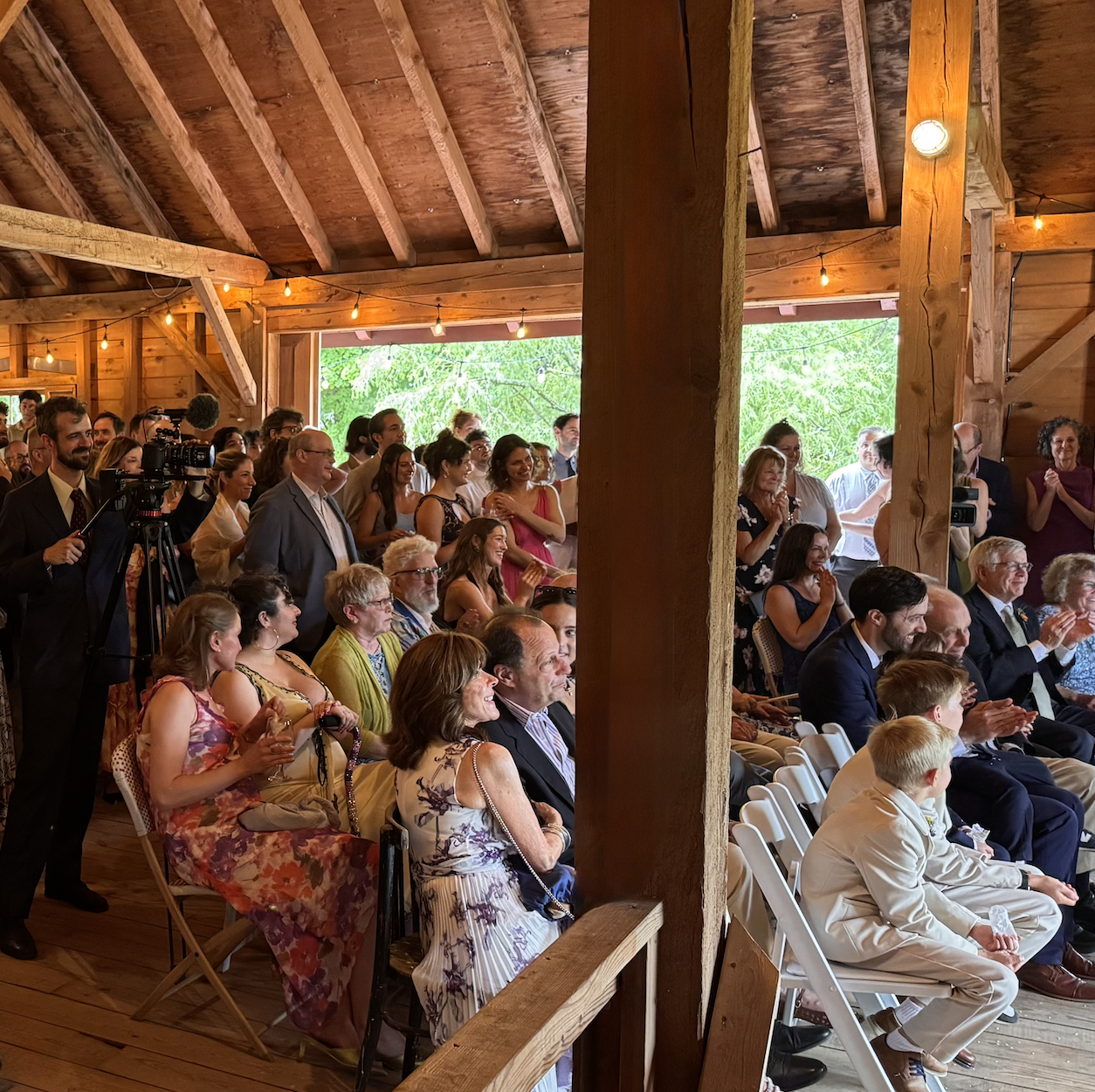 People attending a wedding or ceremony inside a rustic wooden barn, sitting and standing, clapping and smiling, with some filming the event. The barn has exposed beams and string lights, with bright greenery visible outside the open side.