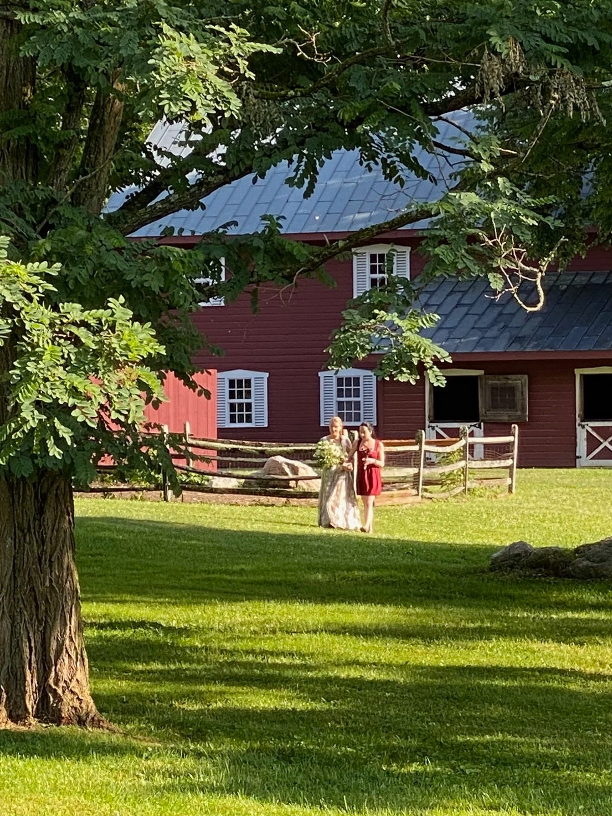 Accompanied by her sister, a bride stands ready on a grassy field near a rustic red barn, surrounded by trees and sunlight.