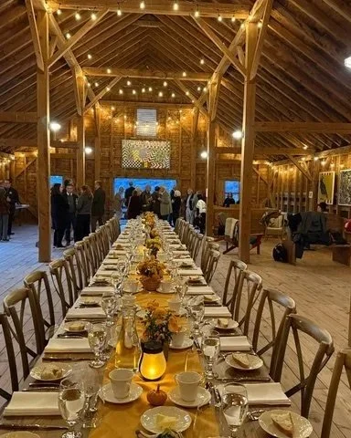Long dining table set with plates, glasses, cups, and flower centerpieces inside a rustic barn with string lights and people socializing.