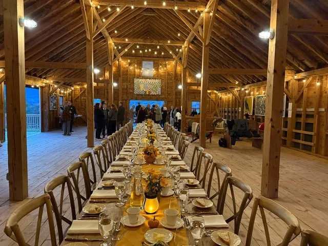 Long dining table set with plates, cups, glasses, and floral centerpieces inside a rustic wooden barn decorated with string lights, with people mingling in the background.
