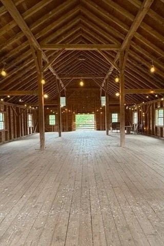 Interior of a large wooden barn with a high, vaulted ceiling, wooden beams, and hanging lights, with open double doors at the far end revealing outdoor greenery.