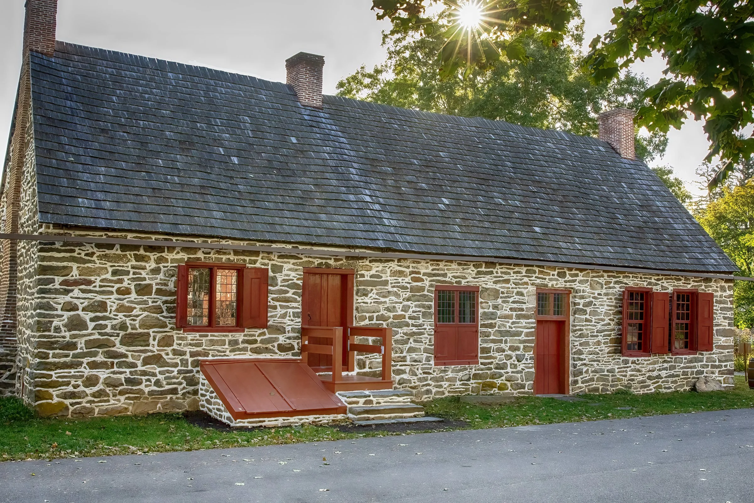 A historic stone house with a dark slate roof, multiple red window shutters, and brick chimneys, with sunlight filtering through a leafy tree in the background.