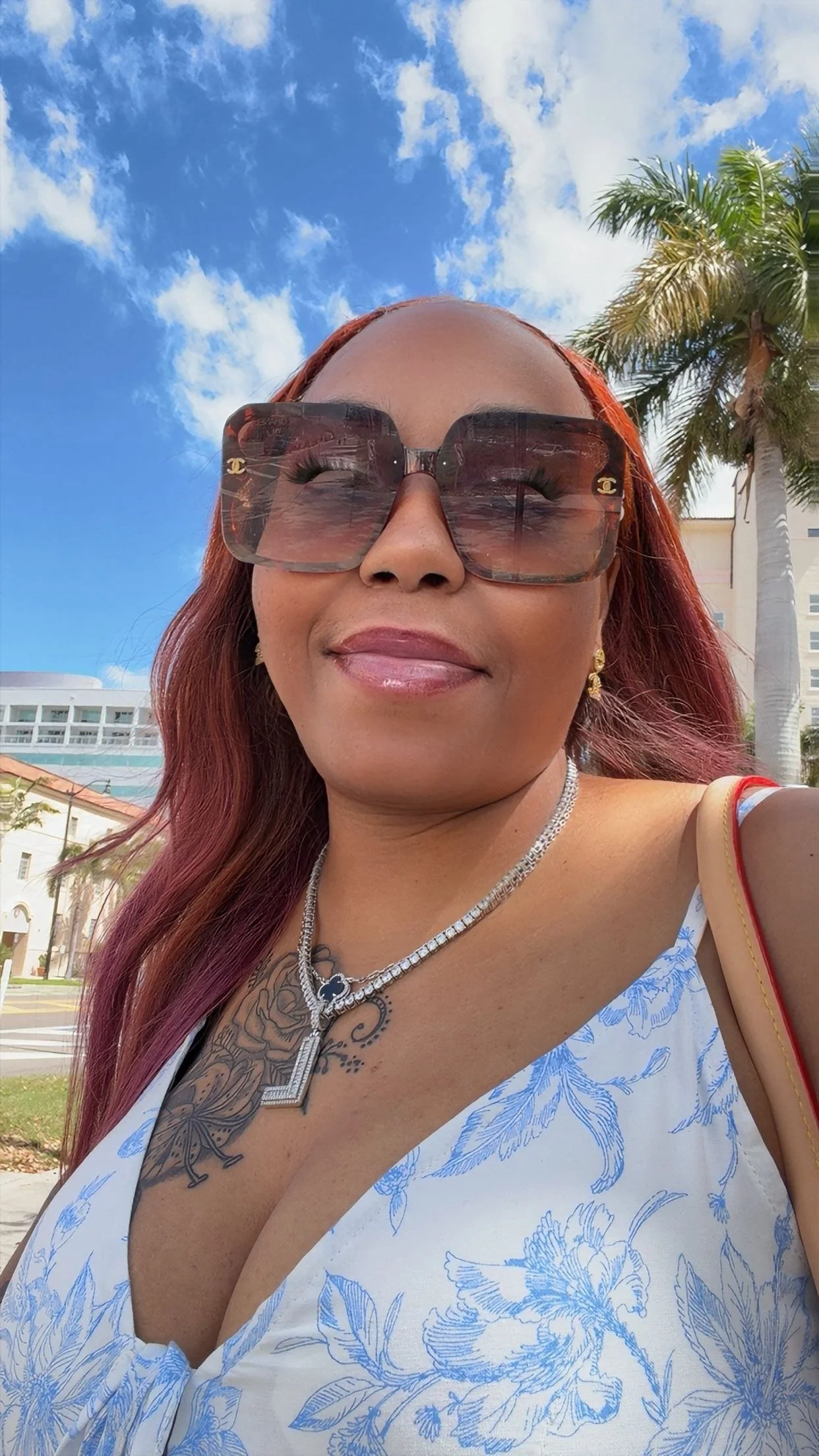 A woman with red hair, wearing large sunglasses with Chanel logos, smiling outdoors on a sunny day. She has tattoos on her chest, is dressed in a white top with blue floral patterns, and is accessorized with layered necklaces and gold earrings. In the background, there are palm trees, a building, and a partly cloudy sky.
