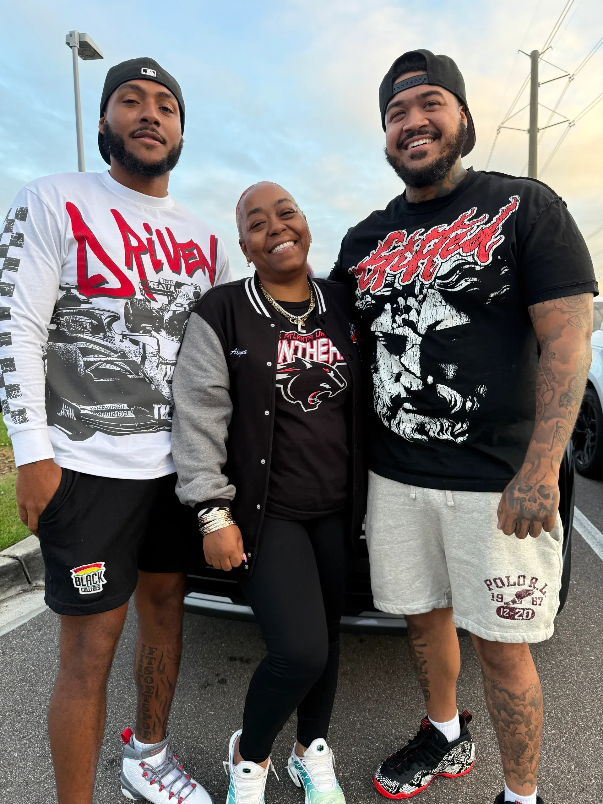 Three smiling people standing outdoors in front of a car and power lines, wearing casual clothes and sneakers.