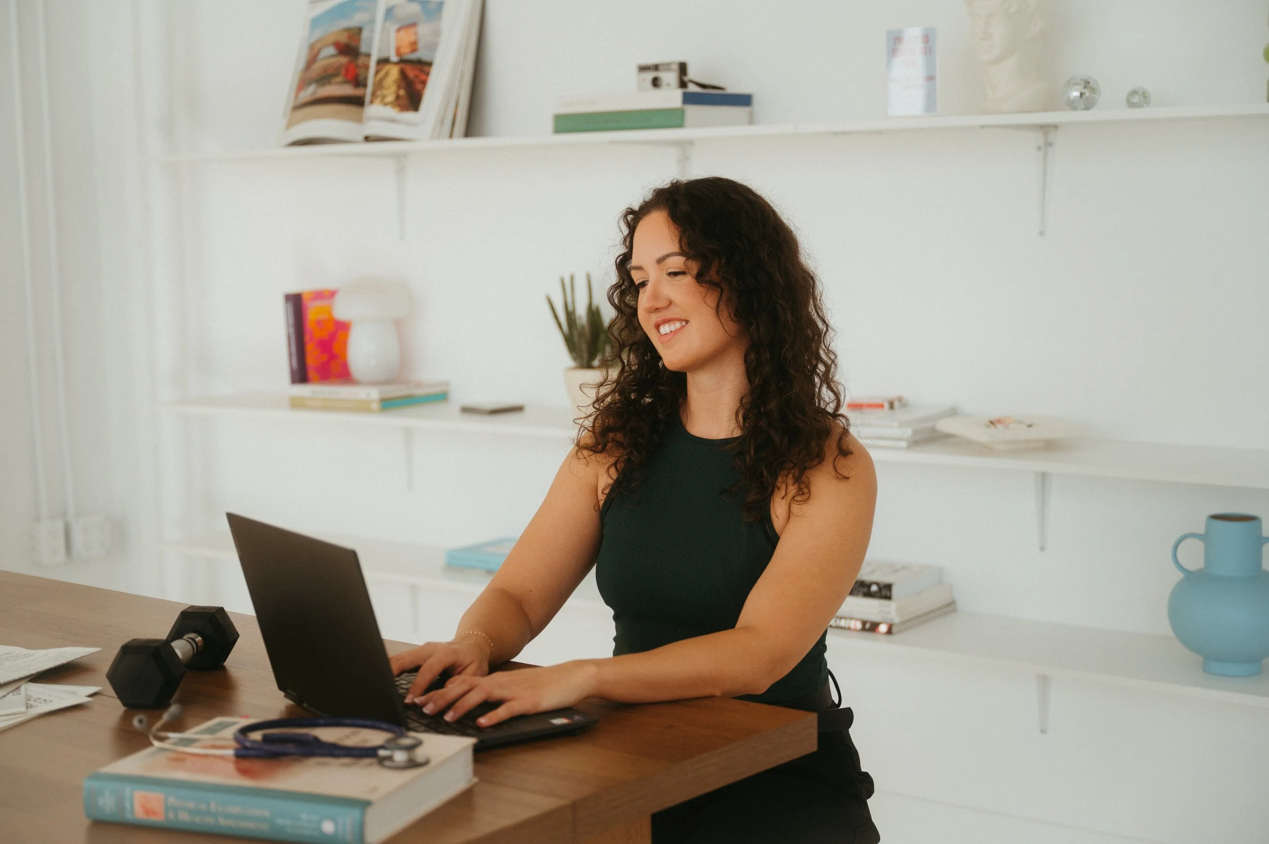A woman with curly dark hair smiling while working on a laptop at a wooden desk in a bright room with white walls and shelves holding books, frames, and decorative items.
