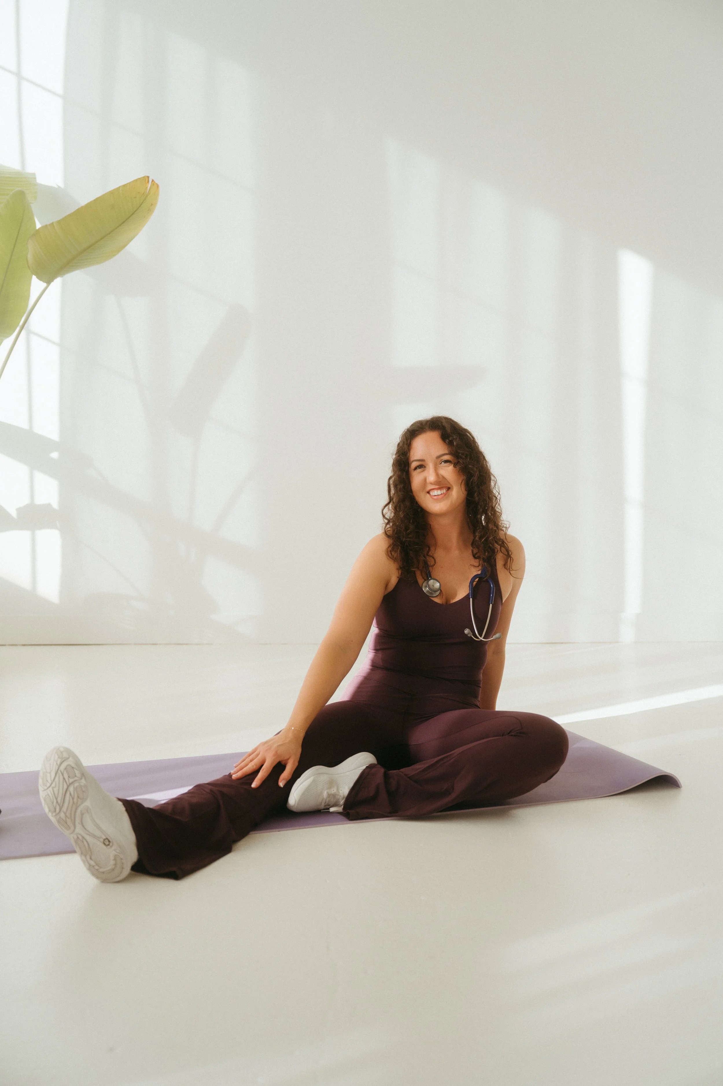 A young woman with curly hair smiling, sitting on a yoga mat, stretching her legs in a bright, minimalistic room with large windows and a potted plant.