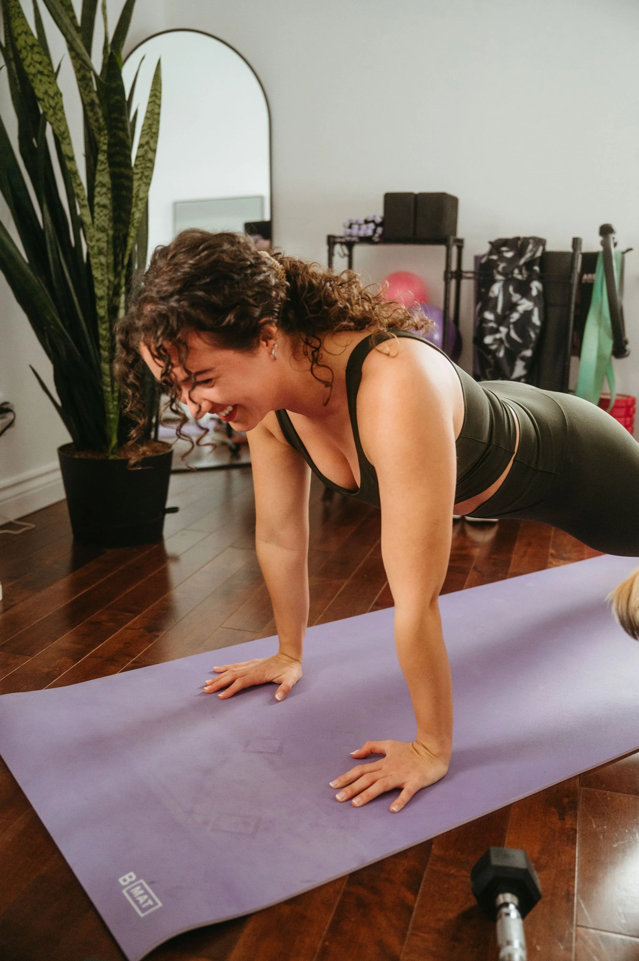 A woman with curly hair smiling while performing a plank exercise on a purple yoga mat in a room with wooden floors, a large plant, and exercise equipment in the background.