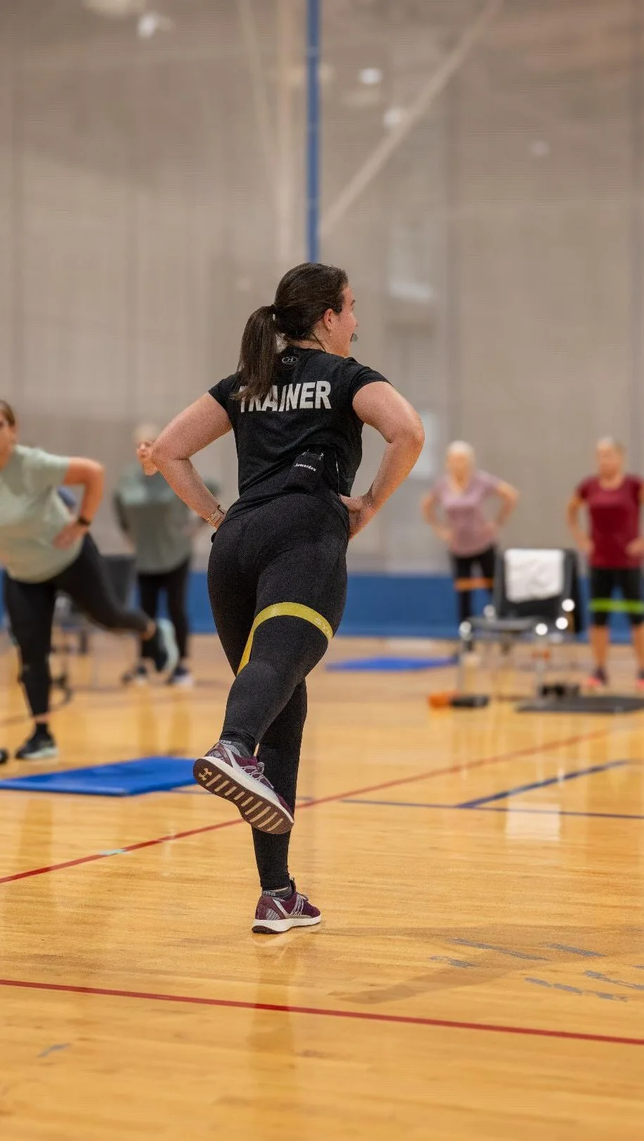 Instructor leading a fitness class in a gym, wearing a black shirt with 'TRAINER' written on the back, and black leggings with yellow accents, with other participants in the background.