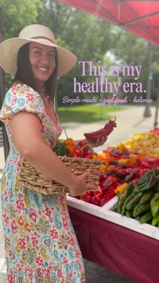 A woman wearing a wide-brimmed hat and a colorful dress holding a basket of vegetables at a farmer's market on a sunny day with green trees in the background.
