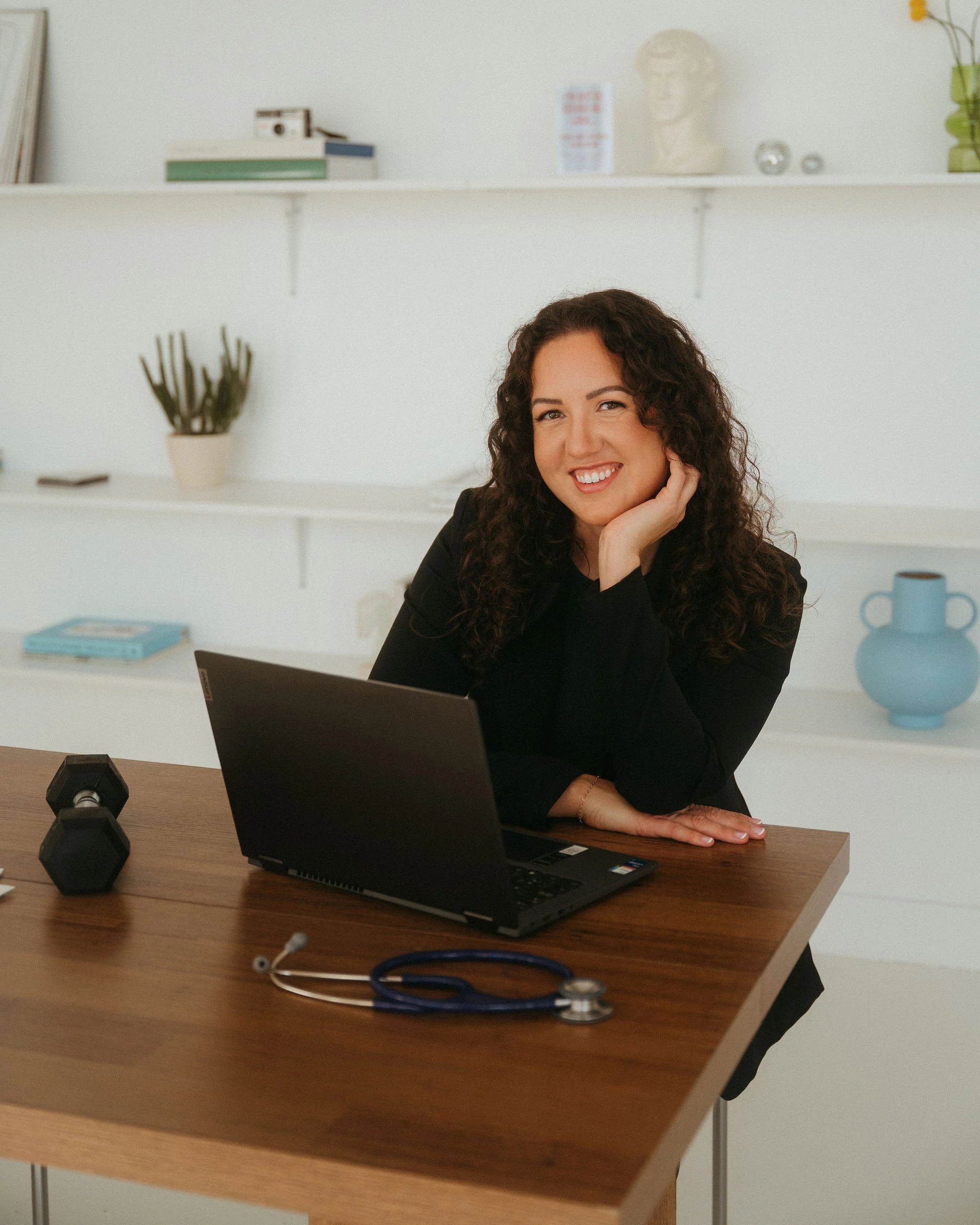 A woman with curly brown hair smiling at the camera while sitting at a wooden desk with a laptop and a stethoscope. In the background, there are white shelves with books, a bust, and decorative objects.