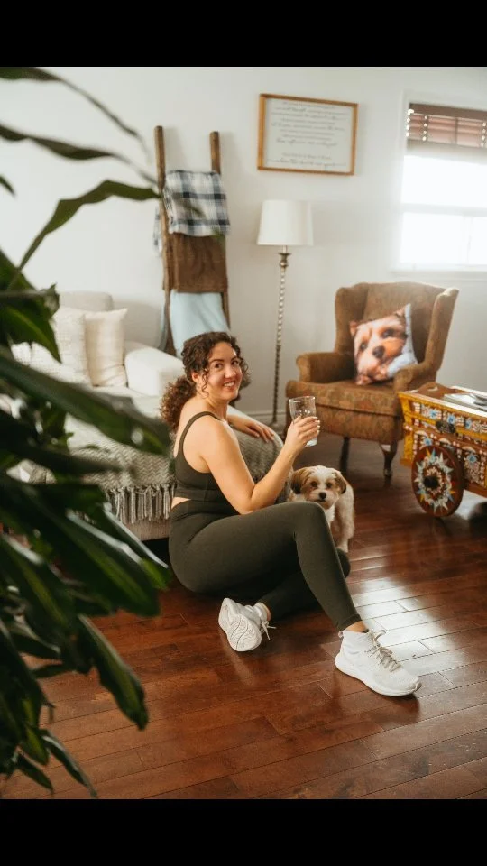 Woman sitting on the wooden floor with a small dog beside her, holding a glass of water, in a cozy living room with armchair, framed picture, side table, and lamp.