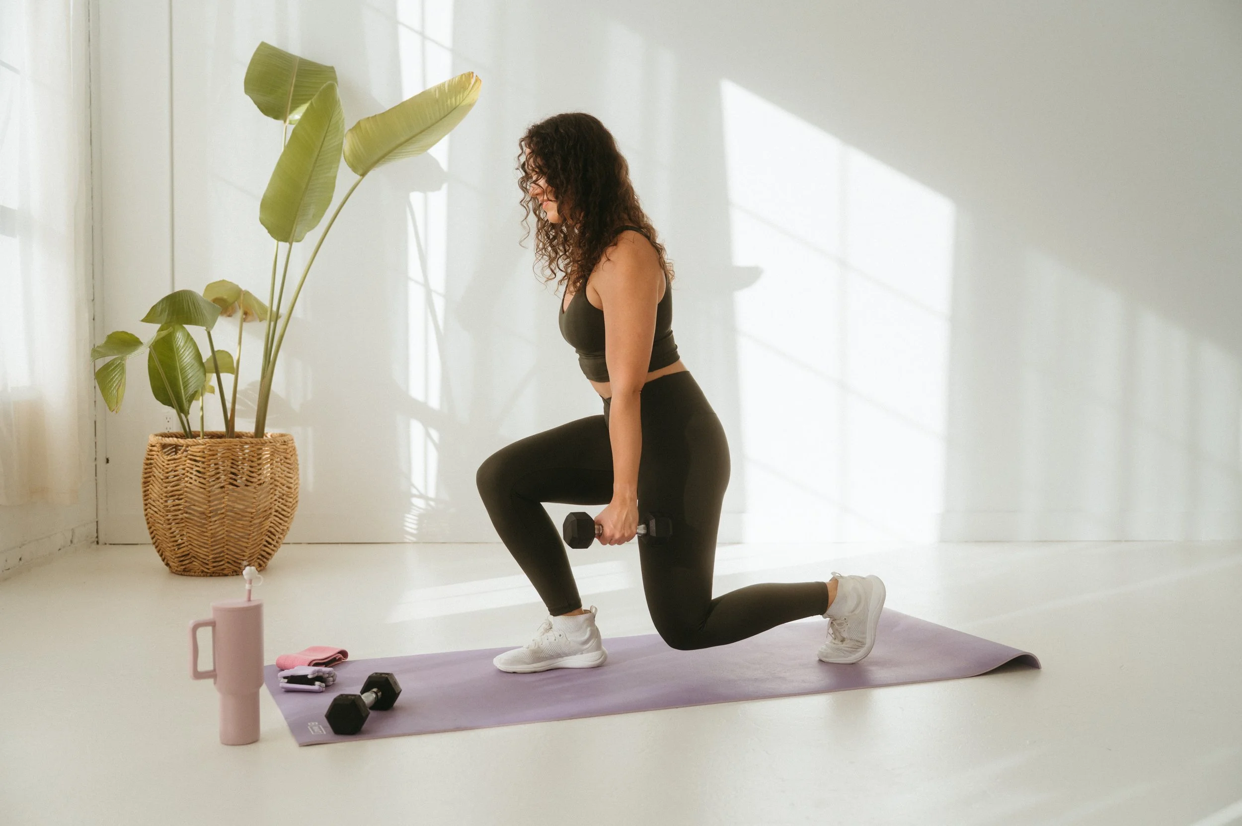A woman in workout attire performing a lunge with dumbbells in each hand on a yoga mat in a bright room with white walls and large windows, with green plants and workout accessories nearby.