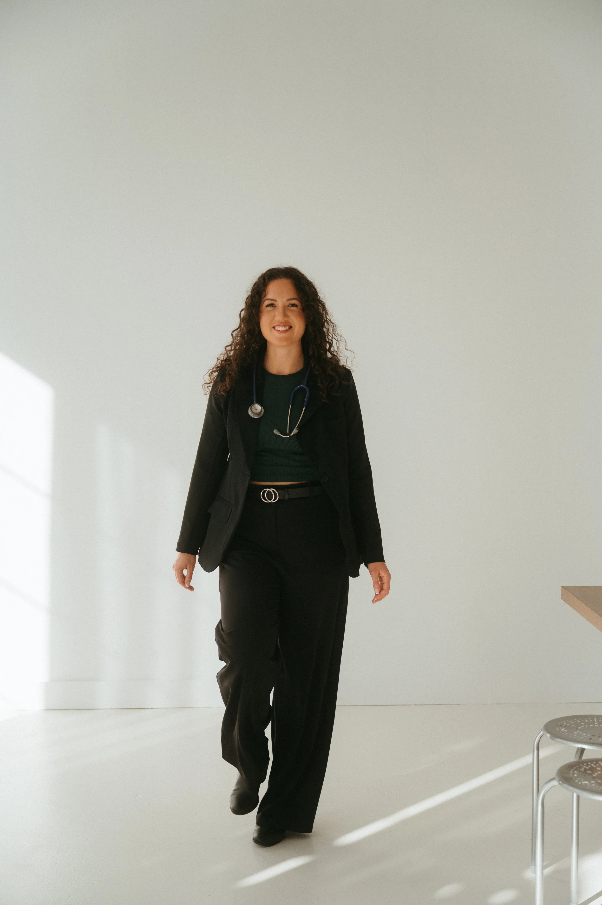 A woman with curly hair, wearing a black blazer, black pants, and a stethoscope around her neck, smiling and walking in a minimalistic room with white walls and natural light.