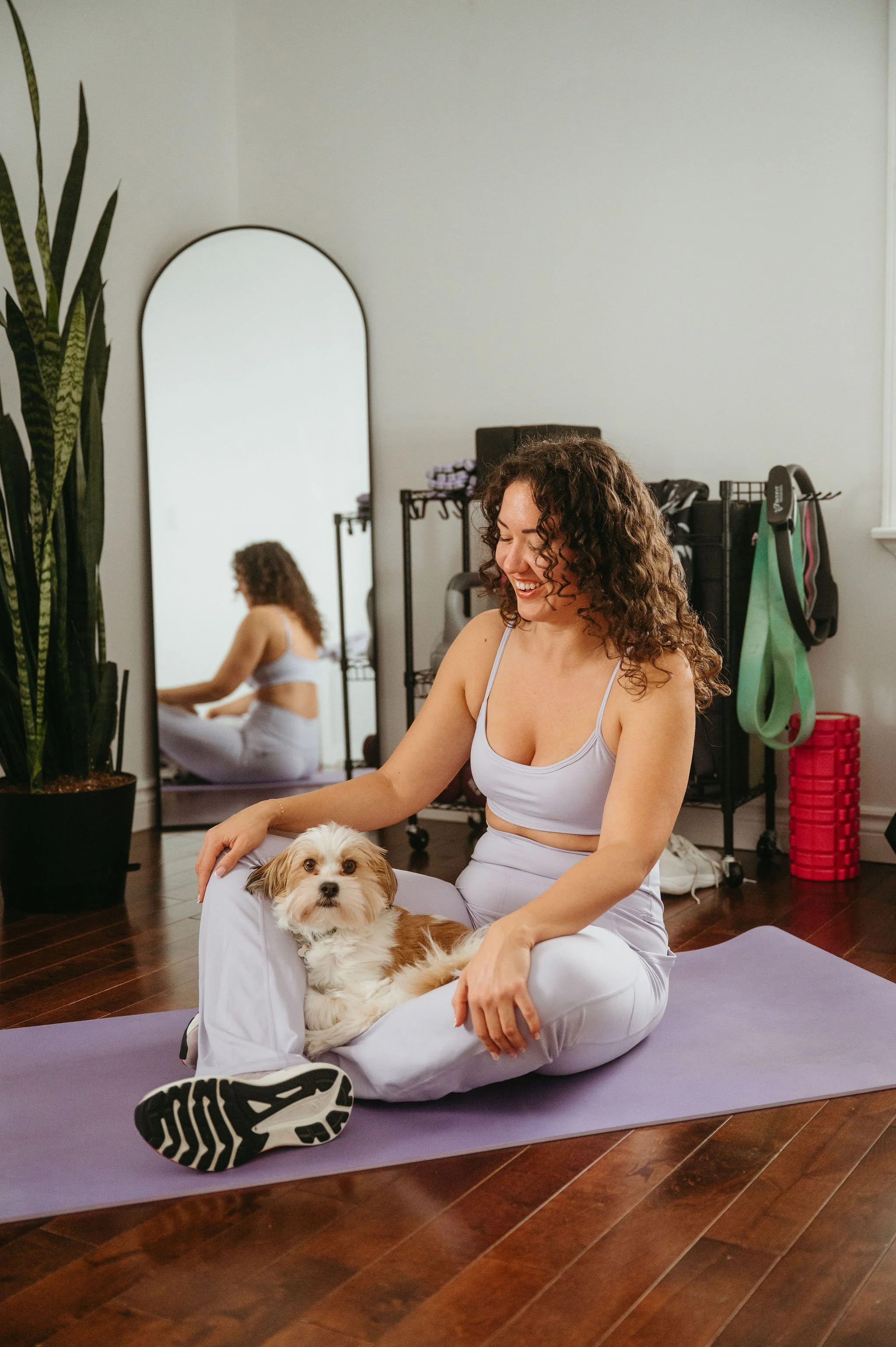 A woman with curly hair sitting on a yoga mat in a room, holding a small dog on her lap. The room has a mirror, exercise equipment, and a ladder-style storage with bags and workout gear. The woman is smiling and looking down at the dog, which looks directly at the camera.