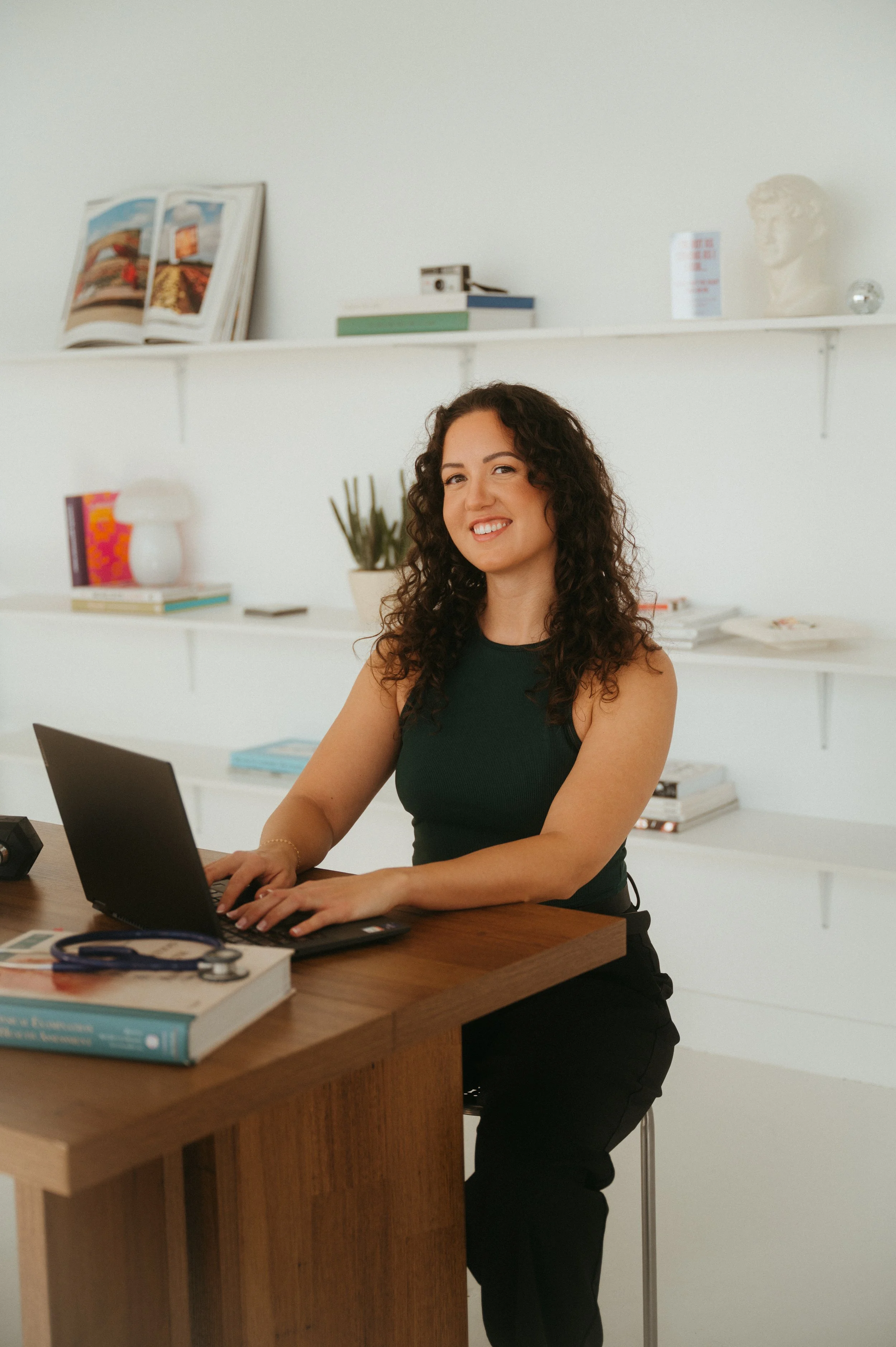 A woman with curly brown hair smiling while working on a laptop at a wooden desk in a bright office. Behind her are white shelves with books, a potted plant, a bust sculpture, and decorative items.