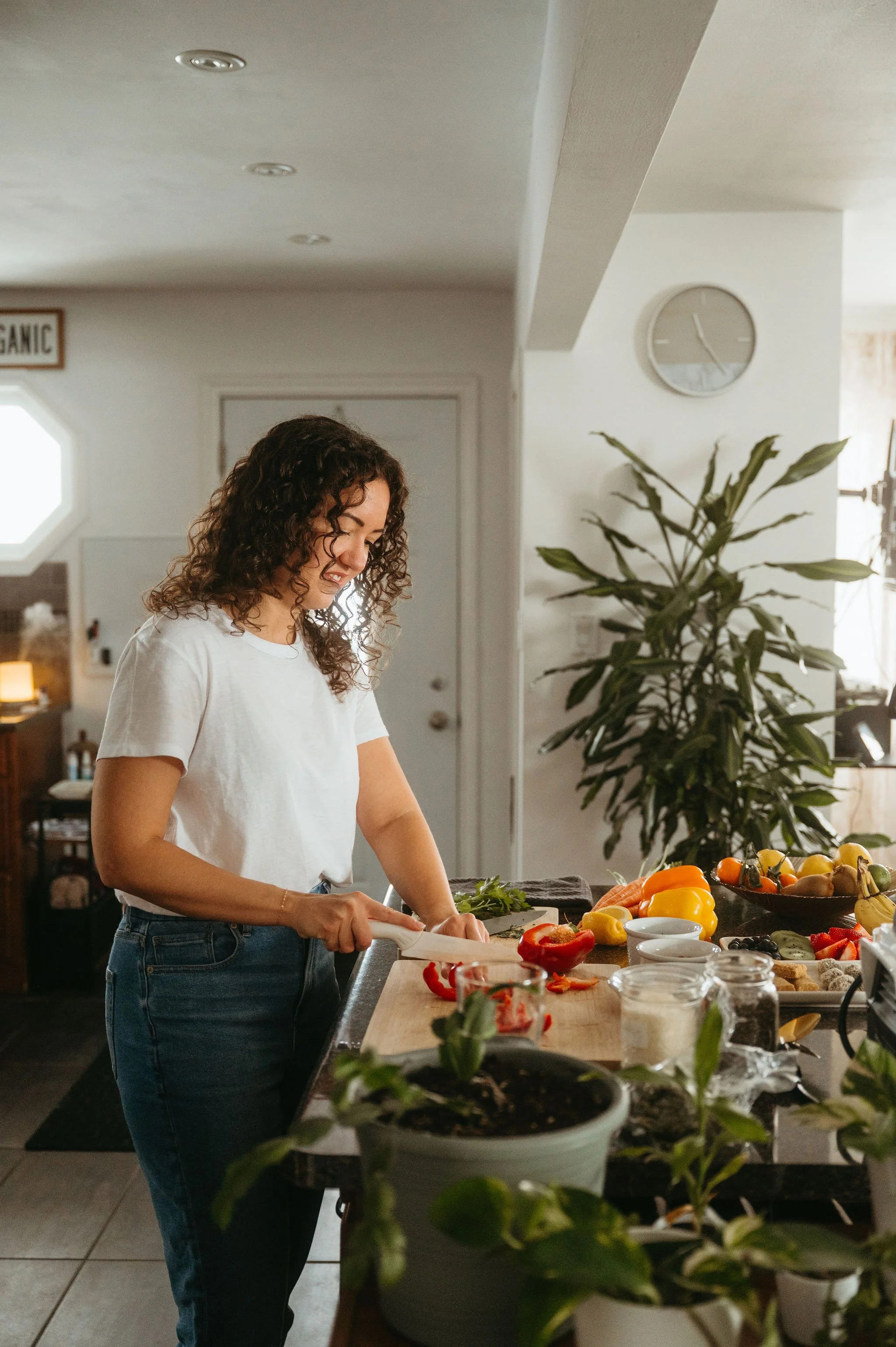 A woman with curly hair preparing vegetables at a kitchen counter. There are various vegetables, bowls, and kitchen items on the counter, with a large potted plant and a wall clock in the background.