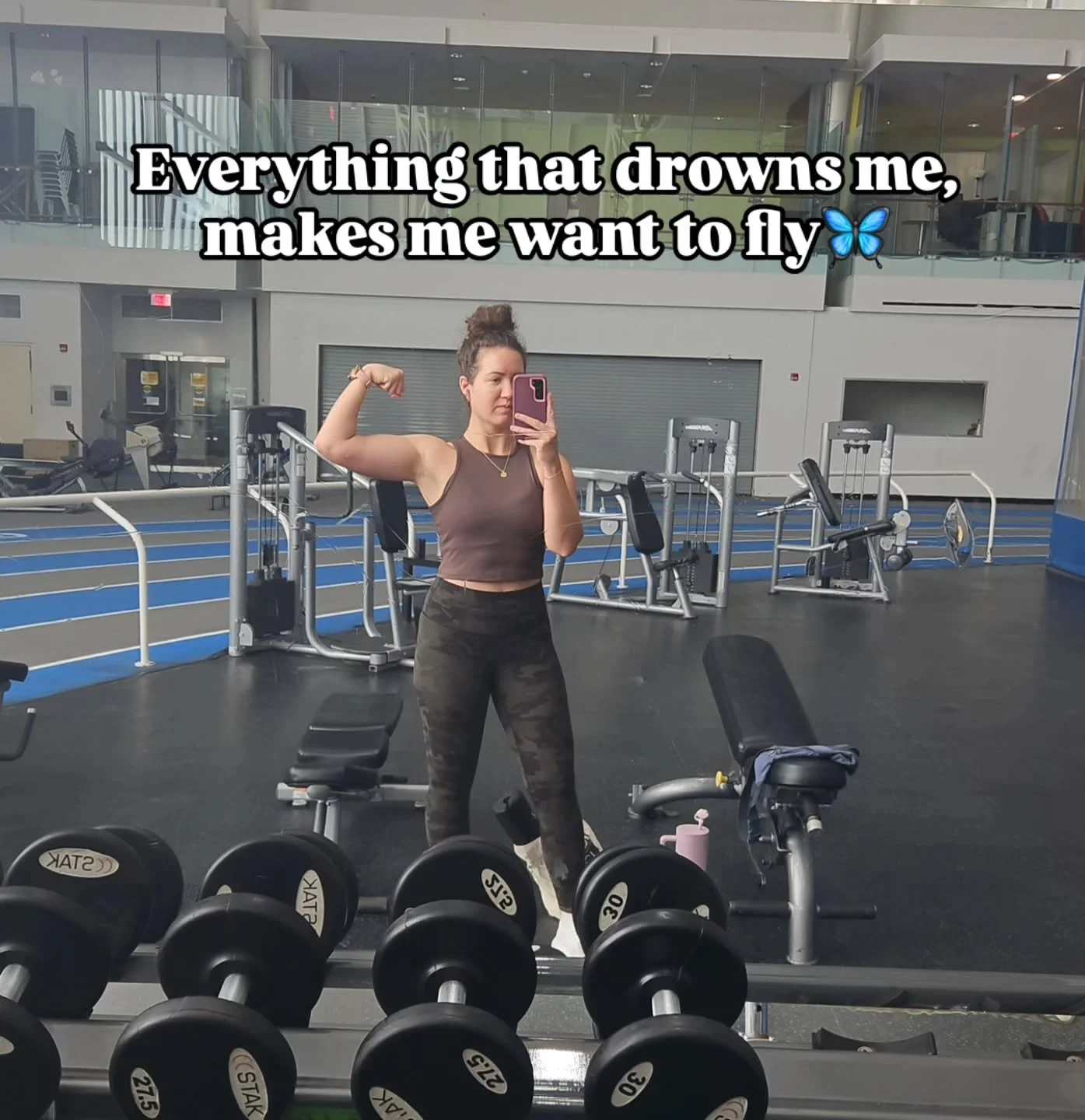 A woman taking a mirror selfie at a gym, flexing her right arm, with weights and gym equipment in the background. The overlay text says, 'Everything that drowns me, makes me want to fly' with a butterfly emoji.