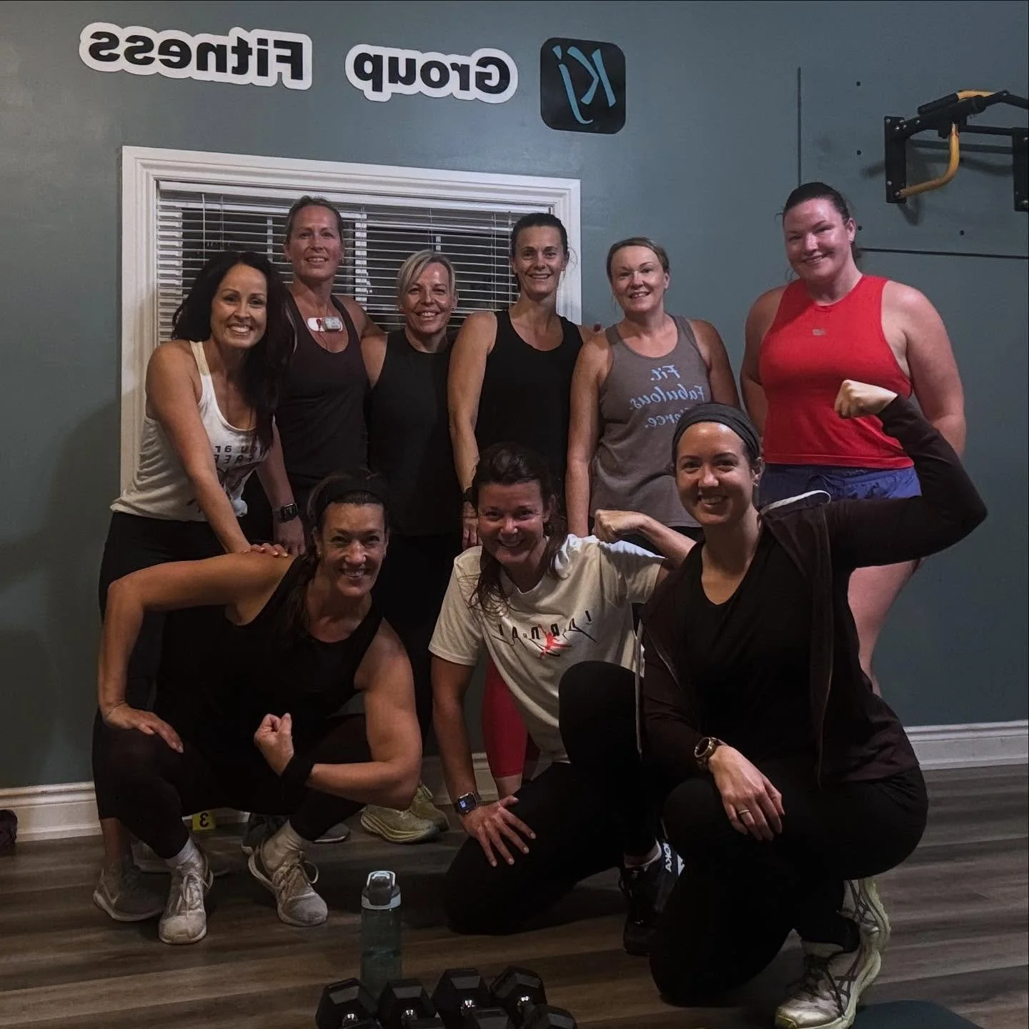 A group of nine women and one man at a gym, posing together indoors with fitness equipment and workout apparel, smiling and showing strength.