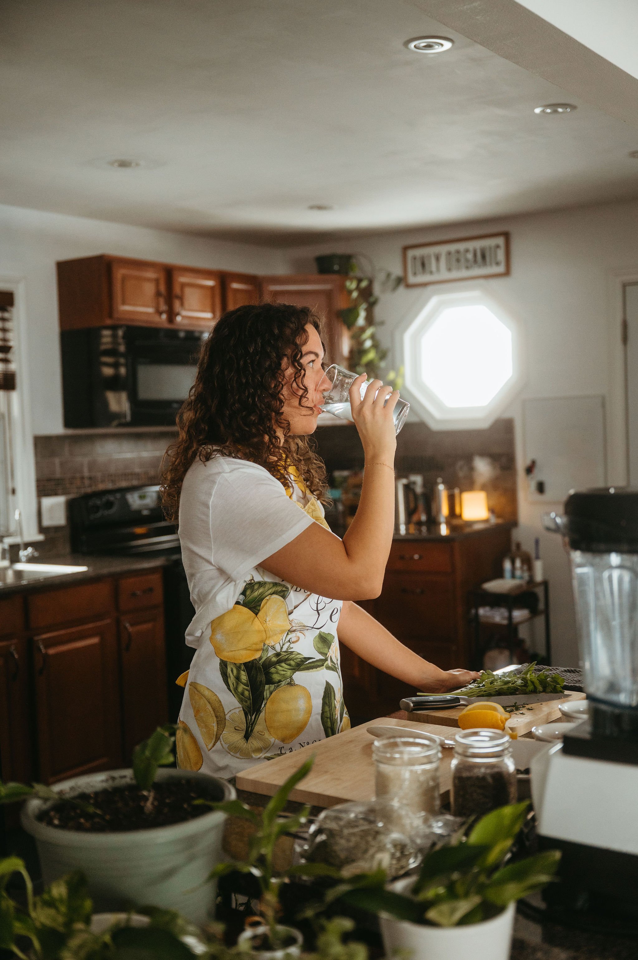 A woman with curly brown hair, wearing a lemon and lime patterned apron, drinks water from a glass in a kitchen. The kitchen has wooden cabinets, a stove, a window with a sign that says 'Enjoy Organic,' and various kitchen utensils and plants on the counter.