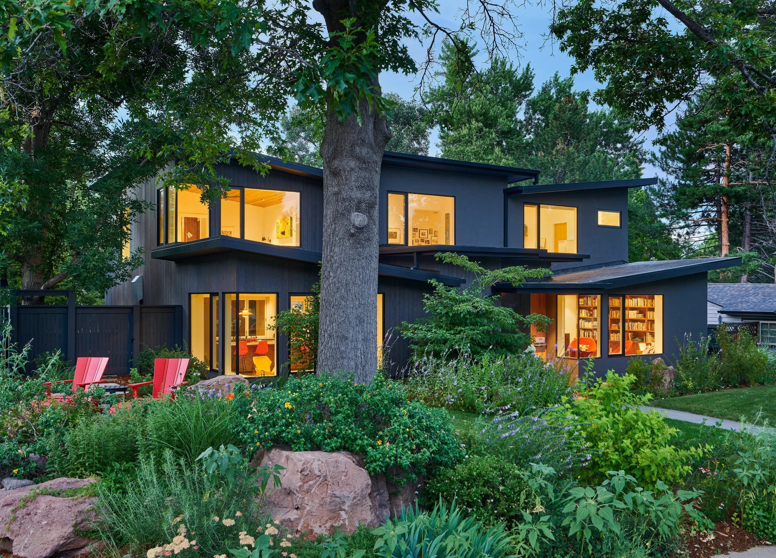 Modern black house surrounded by lush green trees and vibrant garden, with large windows revealing interior lighting at dusk.