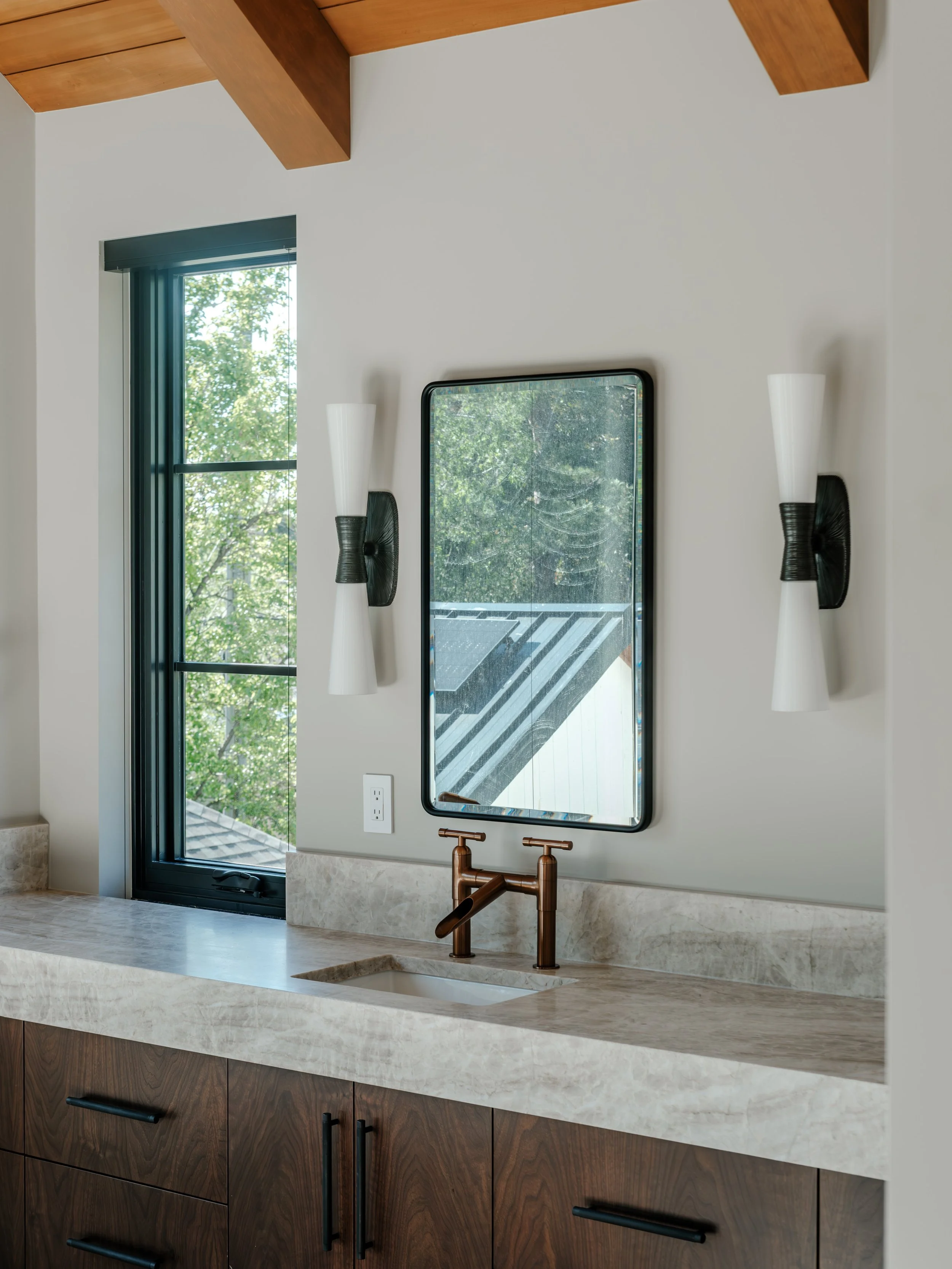 Modern bathroom with wooden cabinets, a white marble countertop, a copper faucet, a large mirror, black wall sconces, and a window with a view of green trees.
