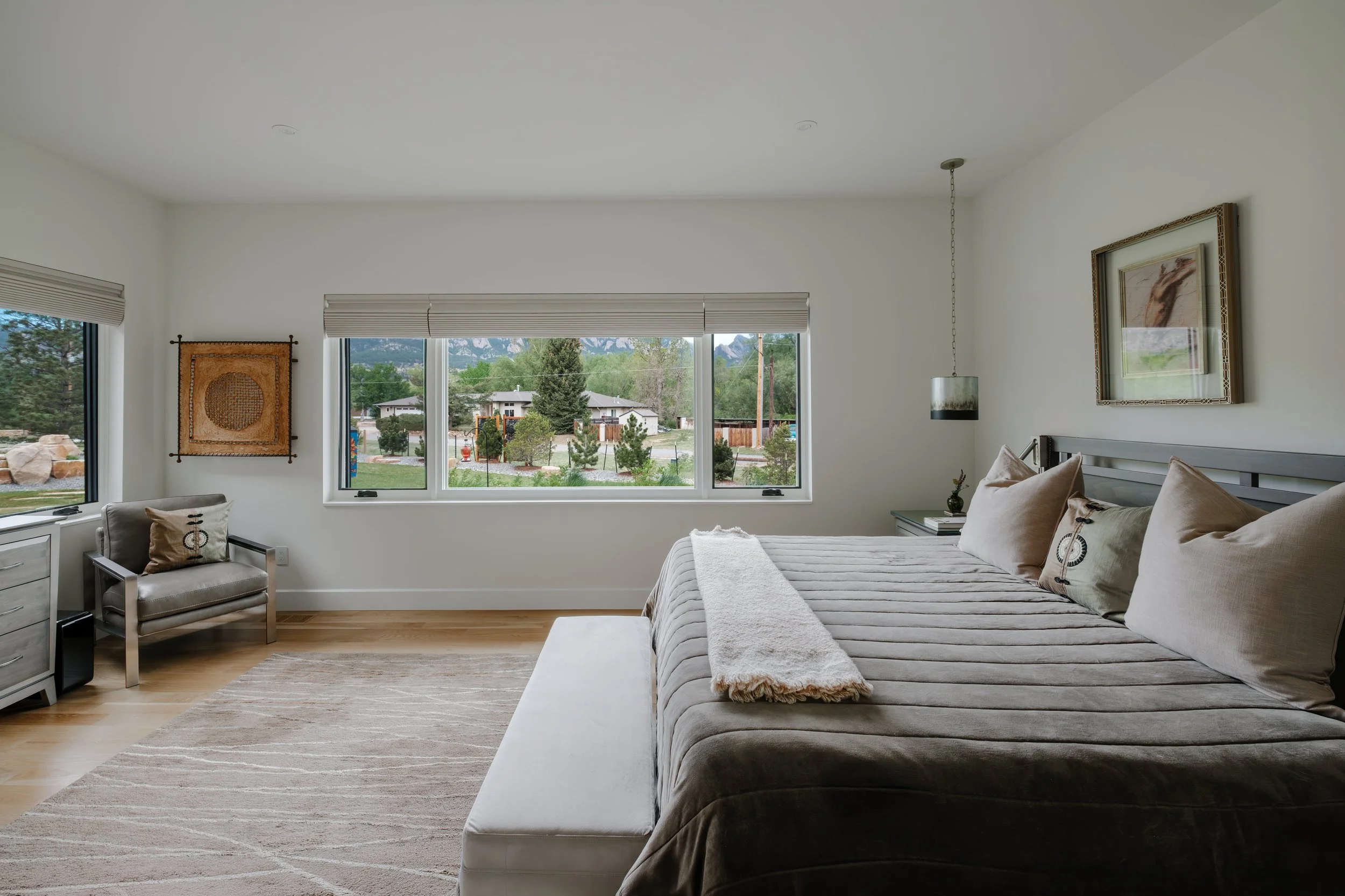 A bedroom with a large gray bed, beige pillows, a white blanket, and artwork on white walls. Large windows show a yard with trees and houses outside.