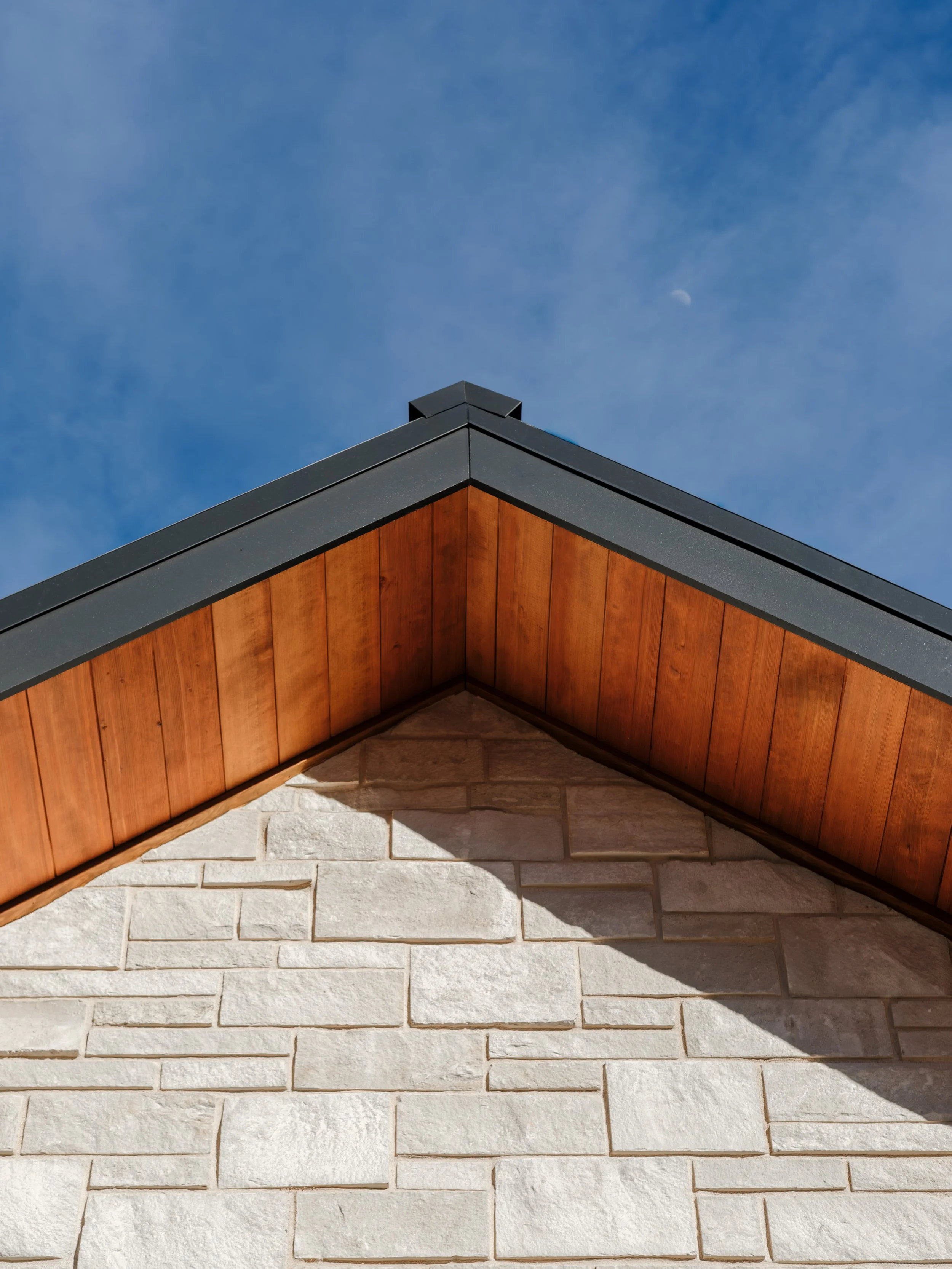 The image shows the corner of a building with a jagged roof, the underside of the roof has wooden paneling, and the wall beneath is made of stone bricks. The sky above is blue with some clouds and a visible moon.