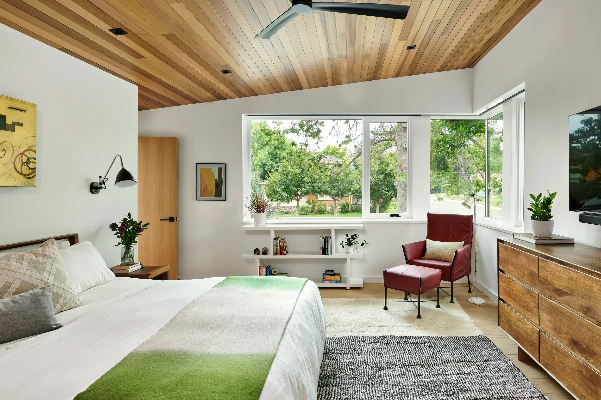 A modern bedroom with a wooden vaulted ceiling, large windows showing green trees outside, a white bed with pillows, a black wall lamp, a framed abstract art, a white bookshelf with books and plants, a red armchair, a wooden dresser, and a black-and-
