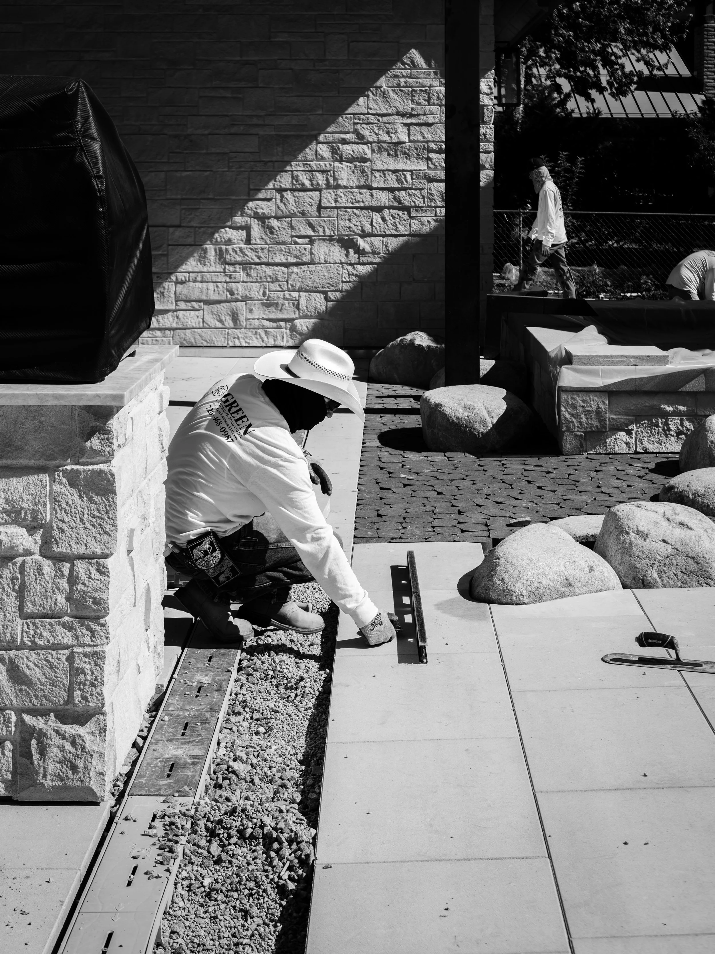 Two workers laying concrete pavers for a sidewalk or patio, with one kneeling and measuring, and another standing nearby, all outside near a stone wall and rocks.