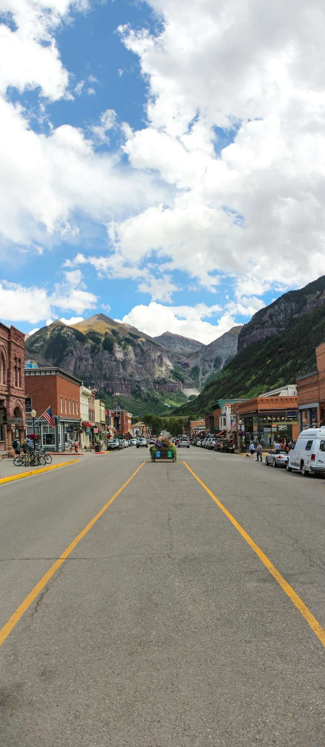 Main street of a small town with shopping buildings on either side, parked cars, bicycles, and pedestrians, with mountainous landscape and partly cloudy sky in the background.