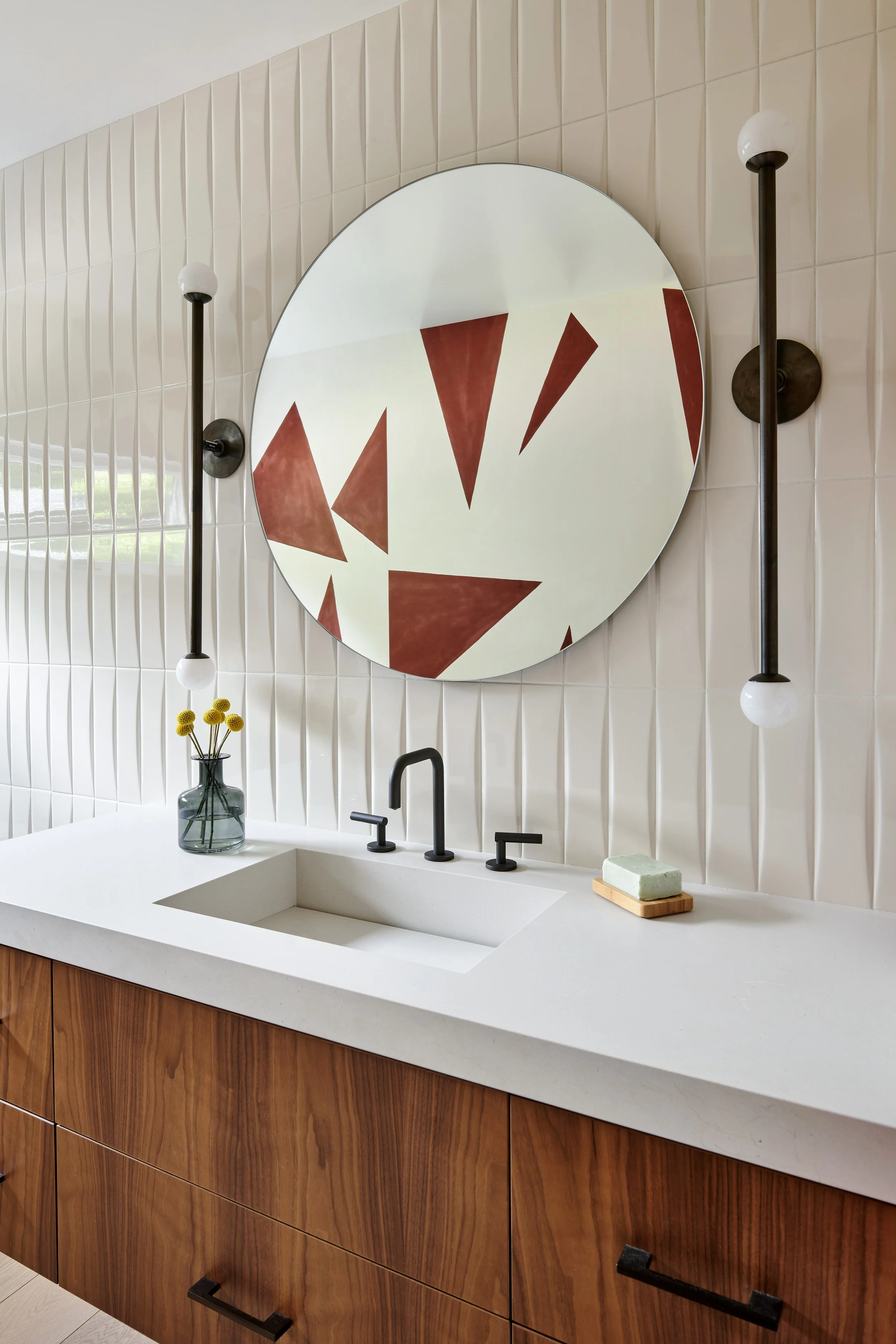 Modern bathroom with a white countertop, black faucet, a vase with yellow flowers, a soap bar on a wooden tray, a large round mirror with geometric brown and white design, and vertical beige tiles on the wall.