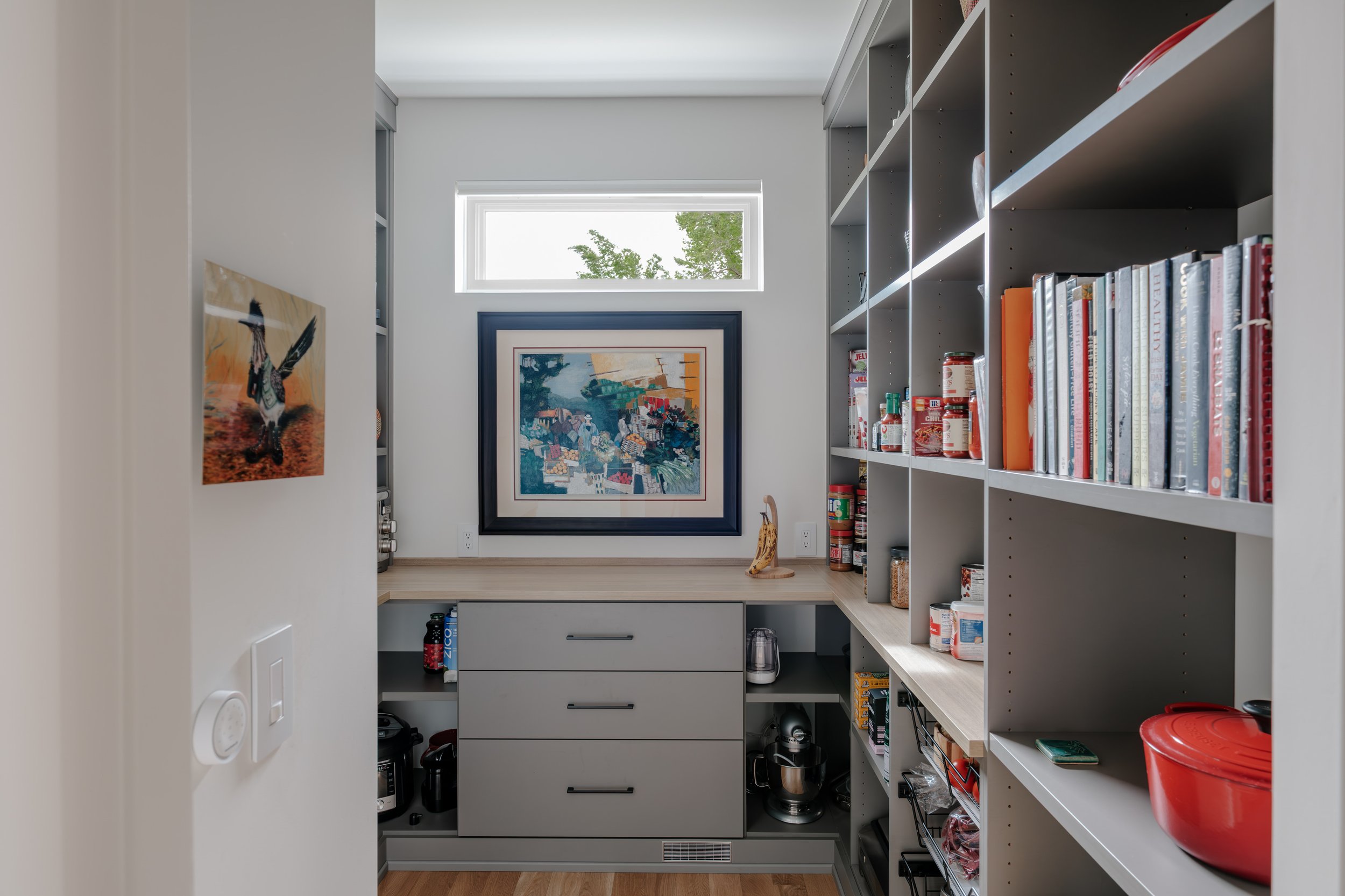 A pantry with white shelves filled with cookbooks and jars, a gray cabinet with drawers, a small window with greenery outside, and colorful artwork on the walls.