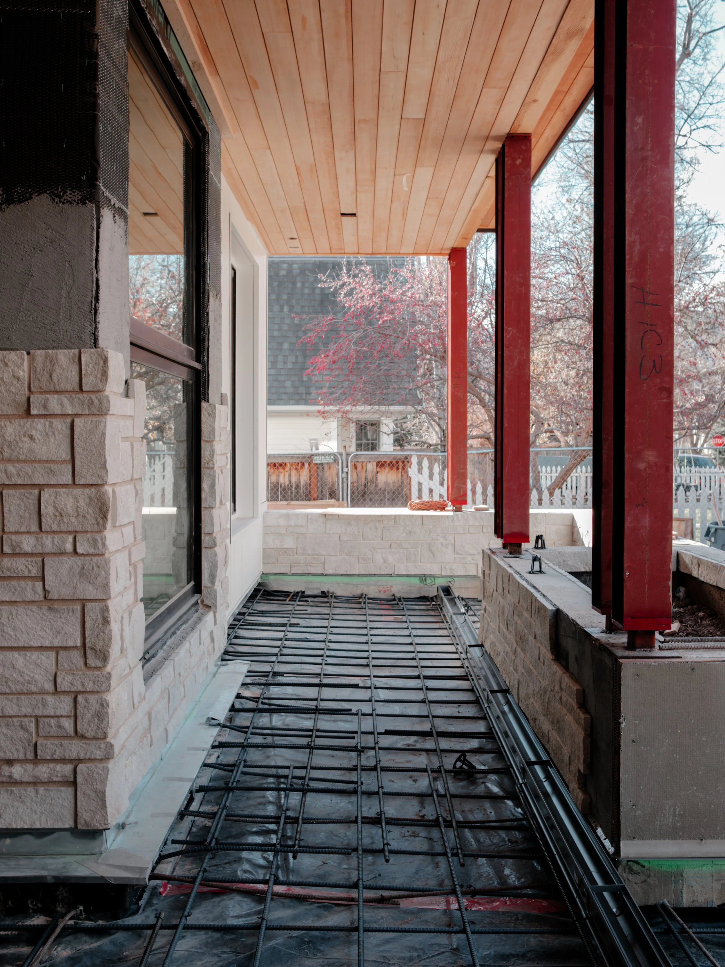 Construction site of a porch or balcony with steel rebar on the floor, brick and stone wall, and red metal support beams, with a wooden ceiling and a view of snowy trees outside.