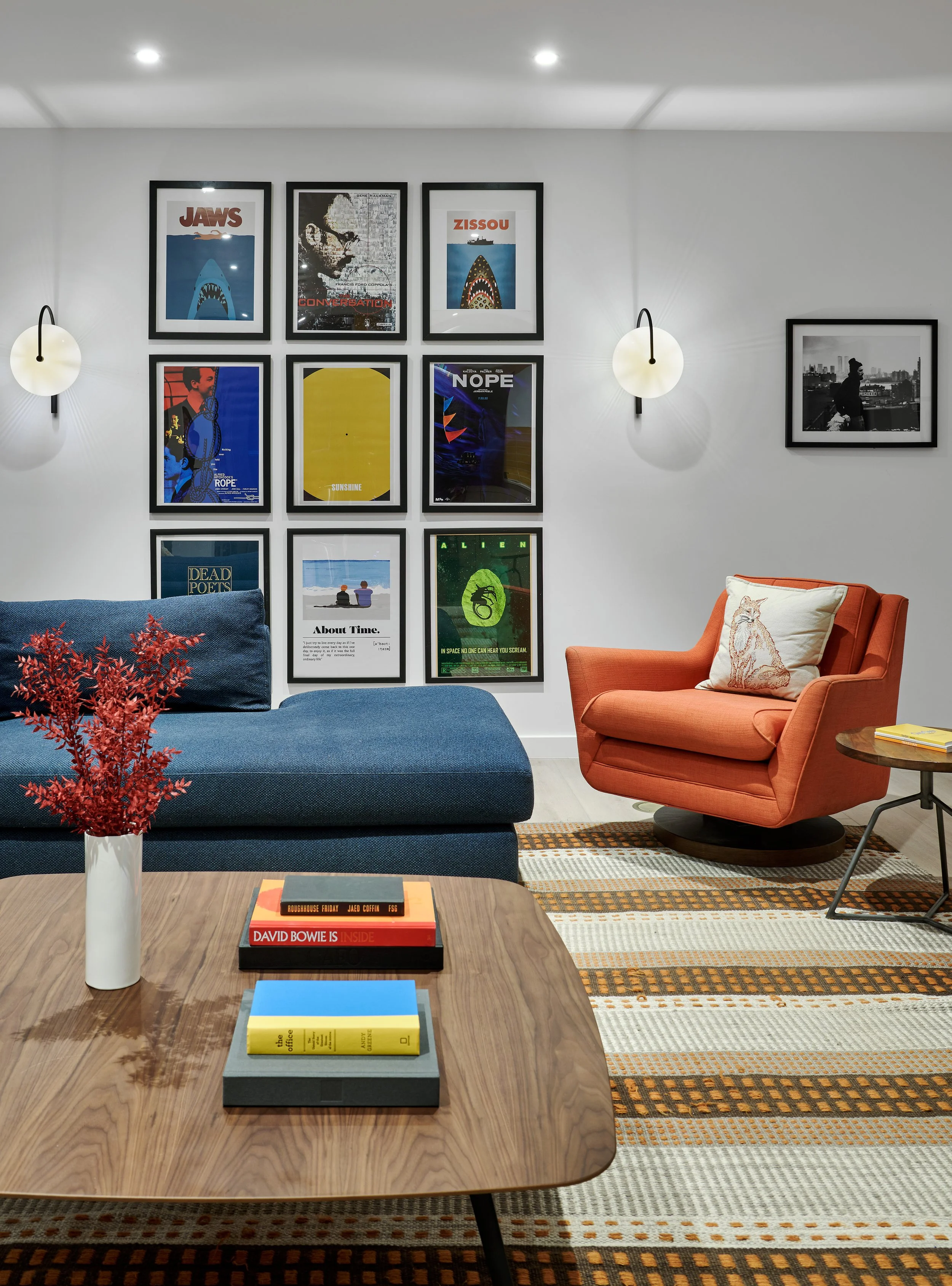 Living room with a wooden table, a blue sofa with a red plant in a white vase, colorful framed artwork on the wall, an orange armchair with a patterned pillow, and a patterned rug.