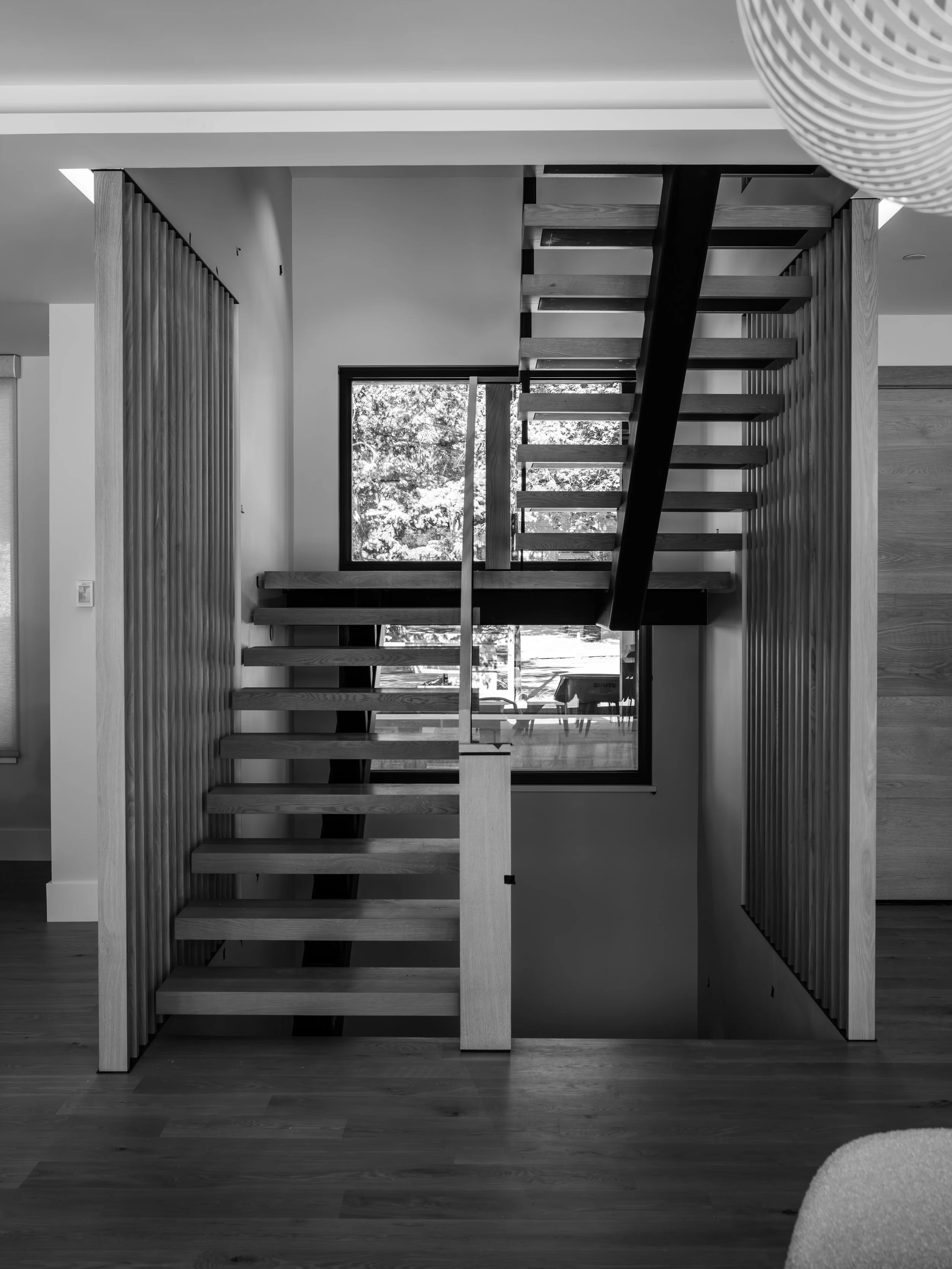 Inside a modern home with a staircase made of wooden steps and black support, viewed from below near a window with trees outside.