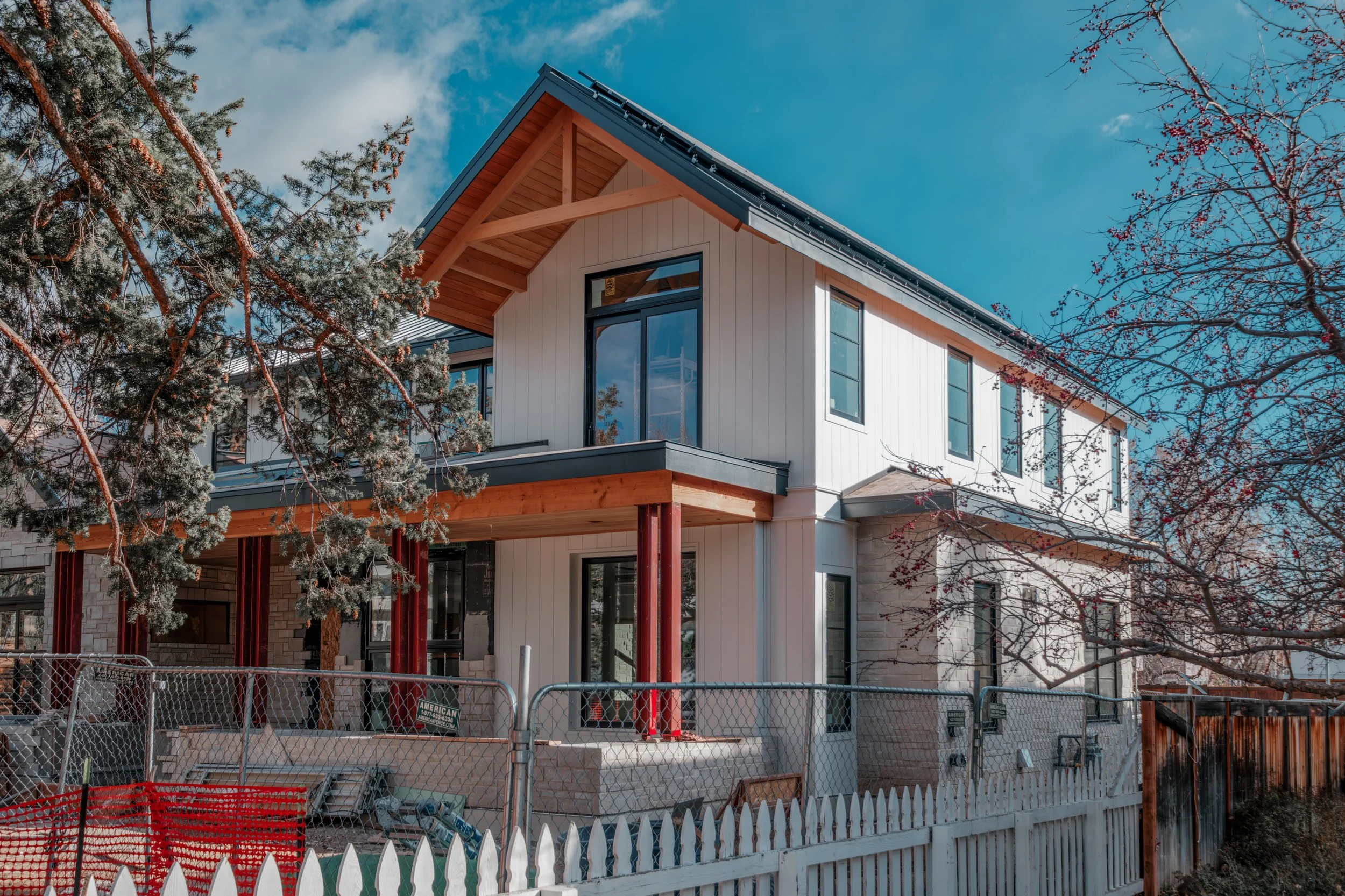 Newly constructed two-story house with a modern design, white vertical siding, large windows, and a gabled roof under construction, surrounded by trees and a construction site with fences.