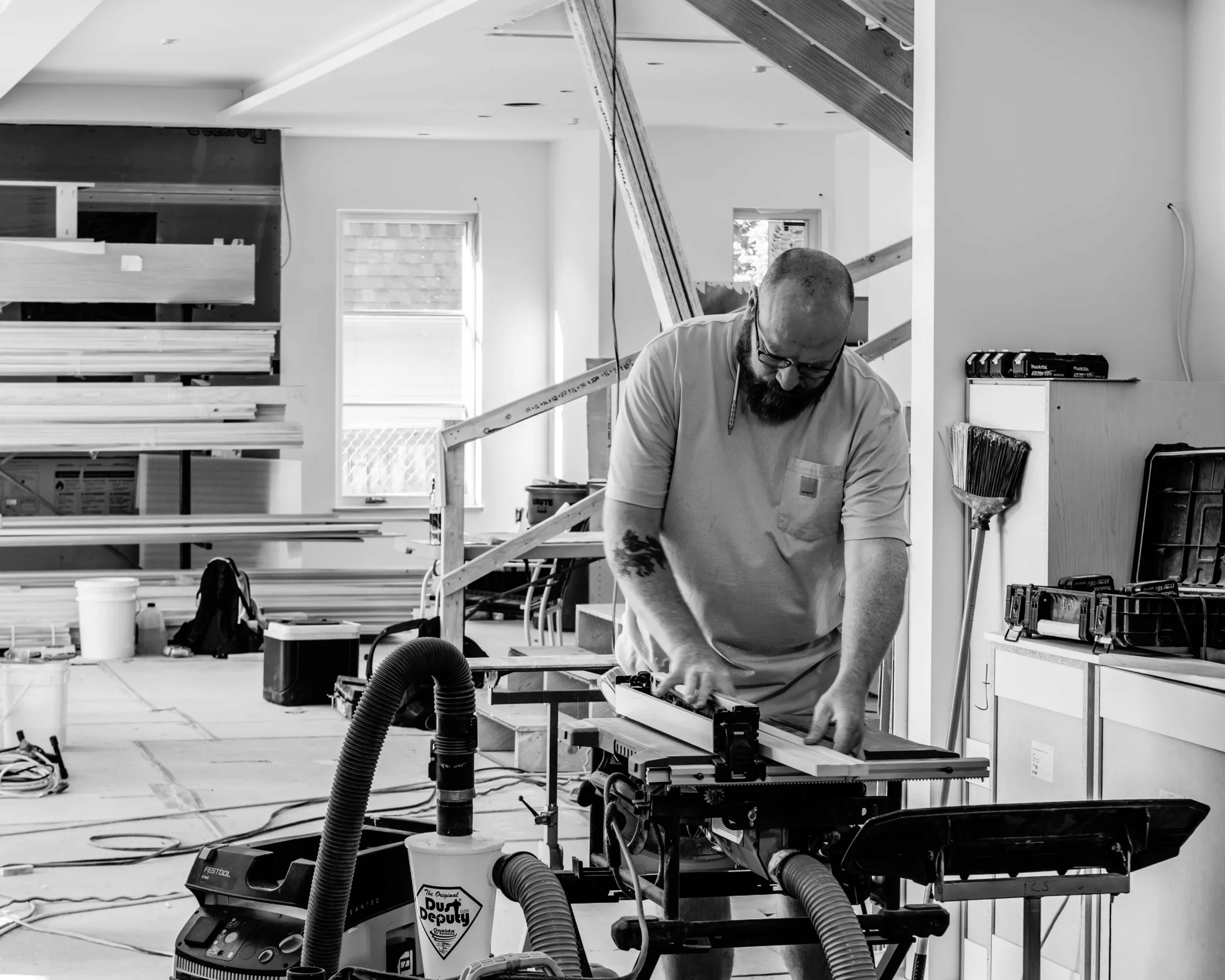 A man with a beard and glasses working in a workshop or construction site, surrounded by tools, wood, and construction materials. He appears focused on a task at a workbench.