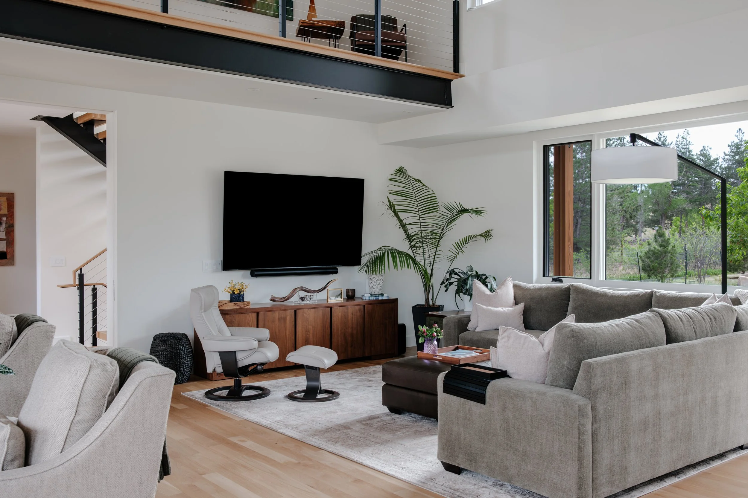 Modern living room with beige sofas, a wooden TV stand, potted plants, a large window with outdoor trees, and a wall-mounted flat-screen TV.